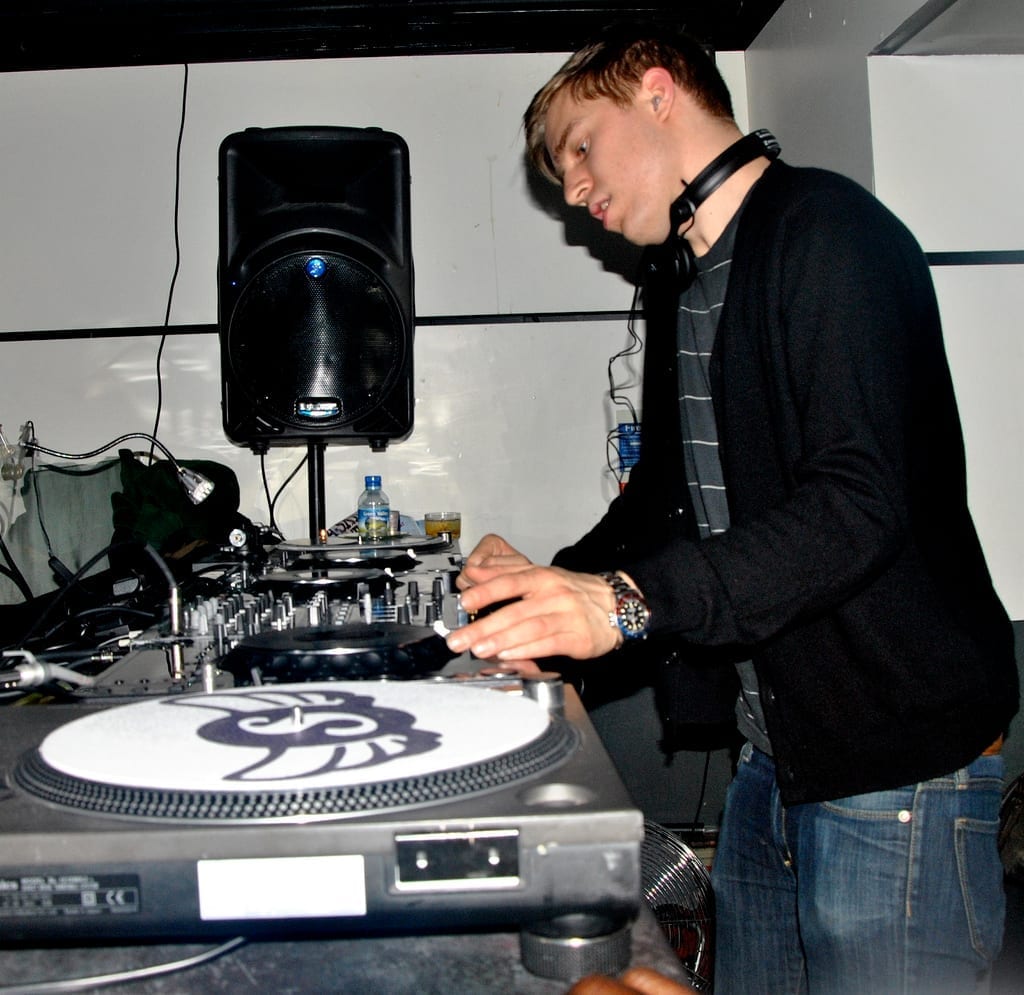 Sub Focus stands at a DJ turntable setup wearing a black cardigan and headphones around his neck, adjusting a mixer.