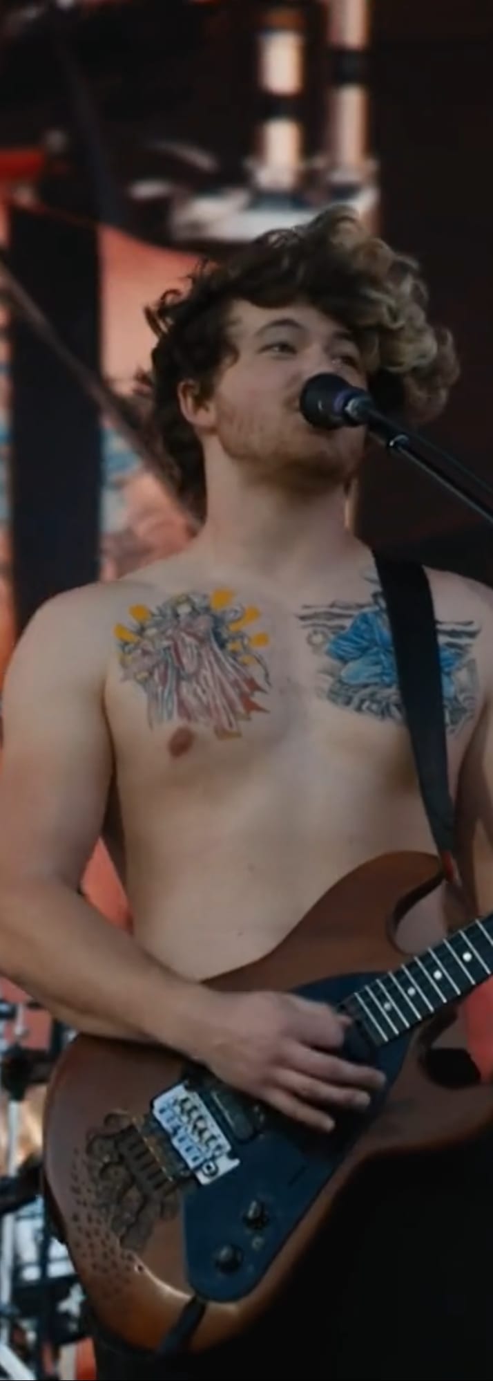 A shirtless, tattooed Sublime band member with curly hair sings into a microphone while playing an electric guitar on stage.