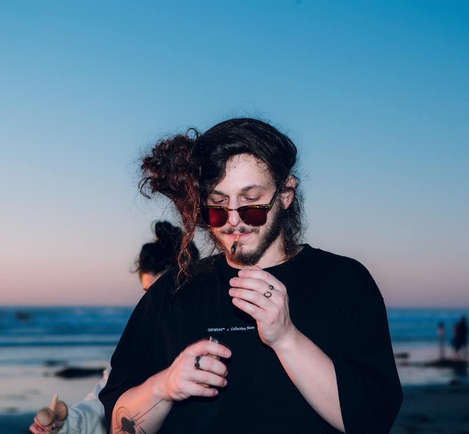 Subtronics, wearing sunglasses and a black t-shirt, stands on a beach at dusk with the ocean and sky behind him.