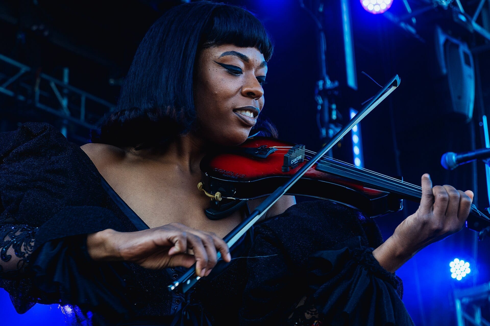 Sudan Archives plays a red electric violin, eyes down and smiling, under blue stage light.