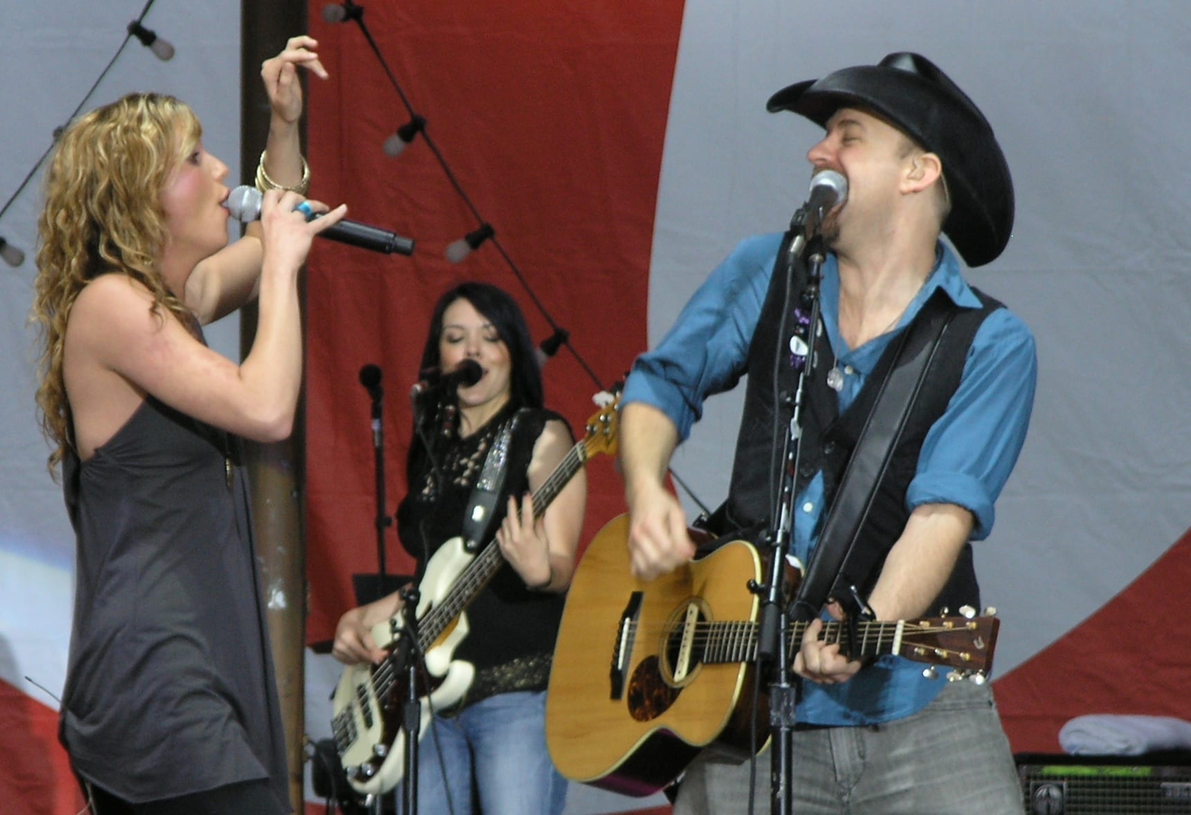 Jennifer Nettles sings into a microphone beside bandmate Kristian Bush, who plays acoustic guitar and sings, with a bassist performing behind them on an outdoor stage.