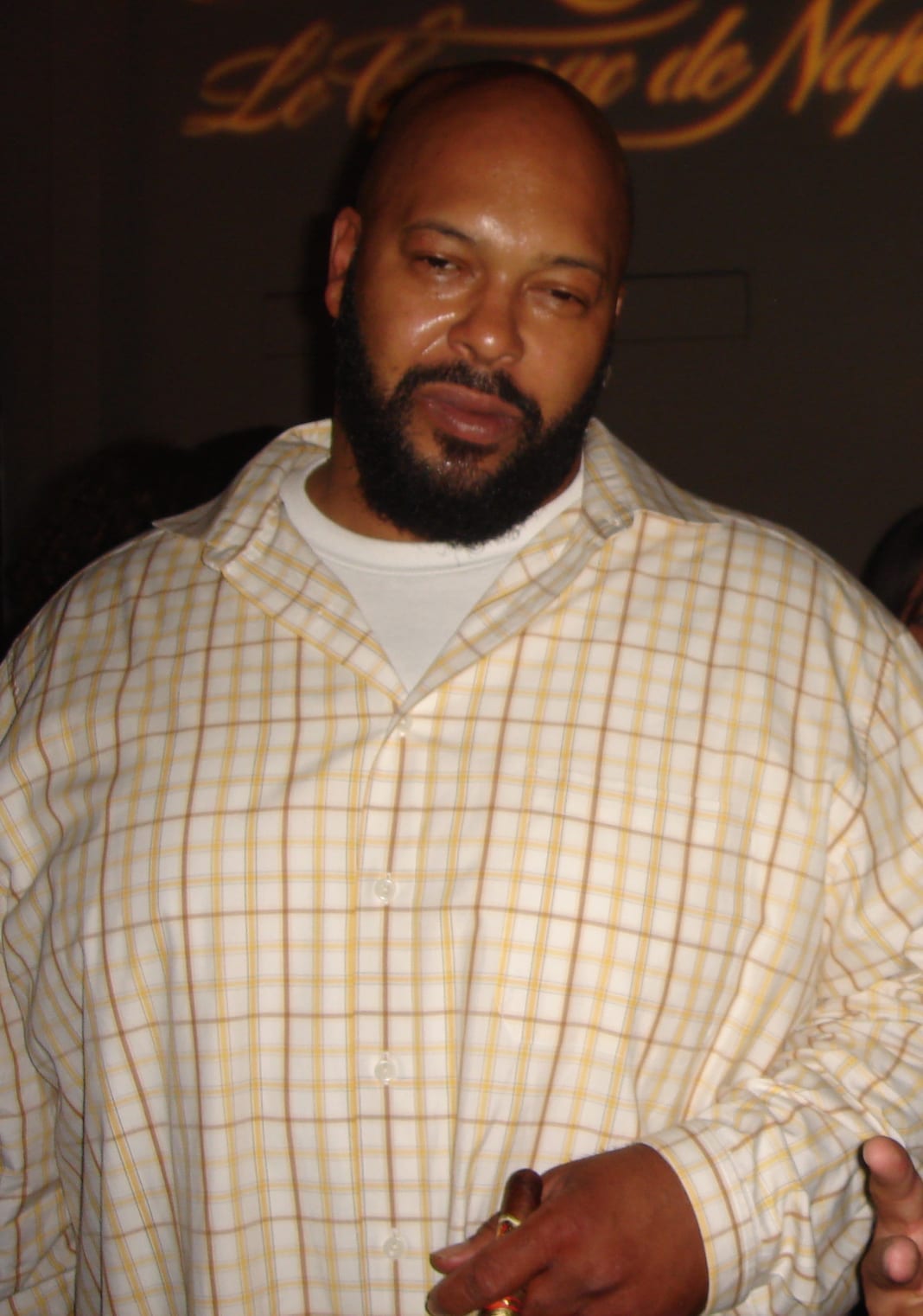 Suge Knight stands indoors wearing a checkered button-up shirt over a white T-shirt, holding a cigar, with a bald head and full beard.