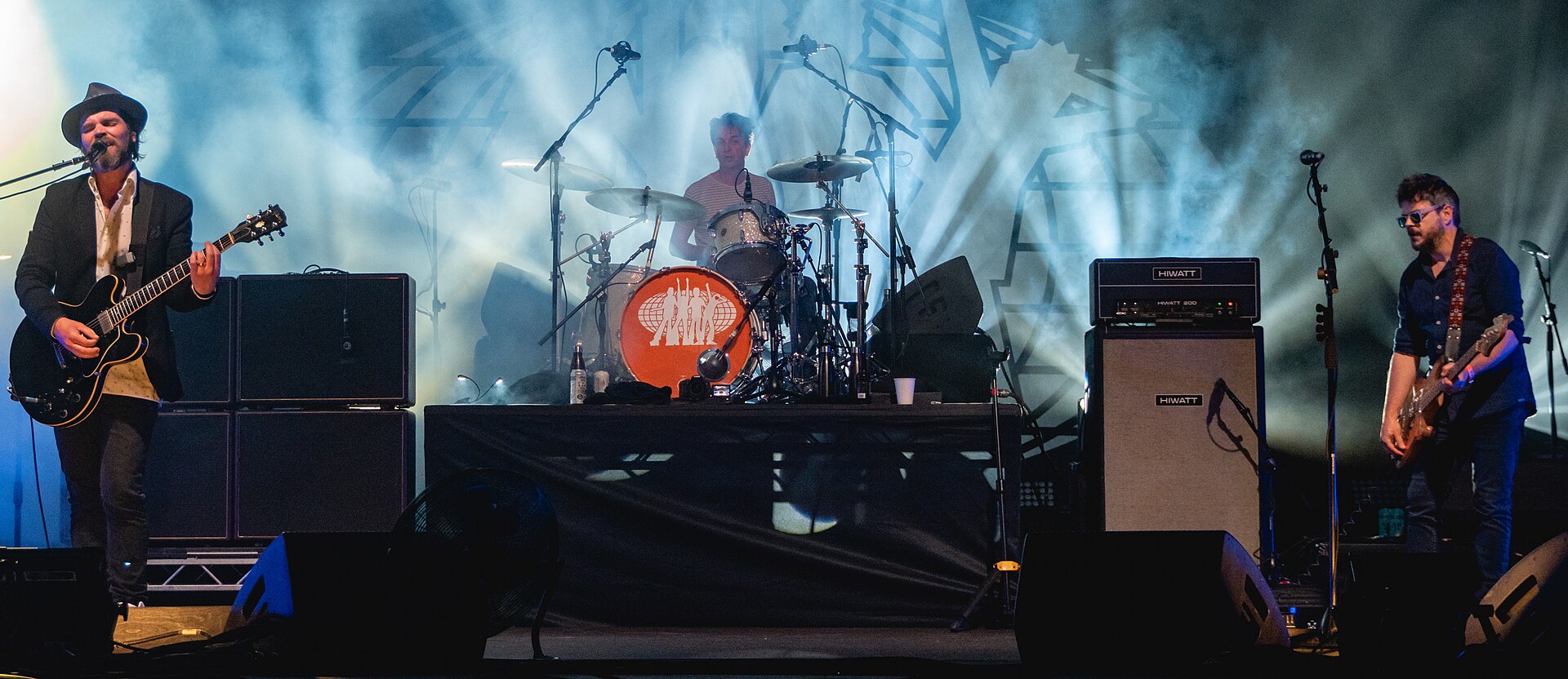 Supergrass perform on an outdoor stage under pale blue light, the singer at a microphone with a guitar, the drummer behind a kit and the bassist at the far right.