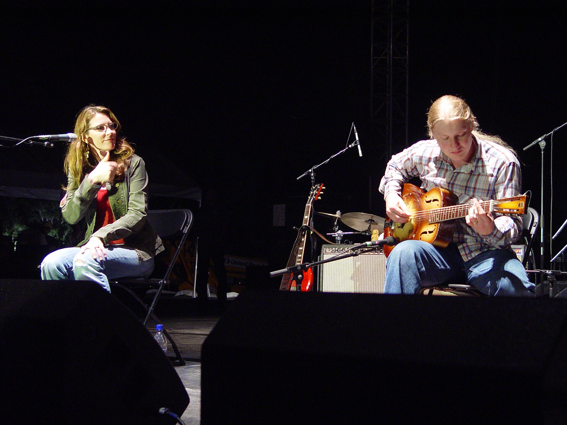 Susan Tedeschi sitting at a microphone on a darkened stage at left while Derek Trucks plays a resonator guitar seated at right