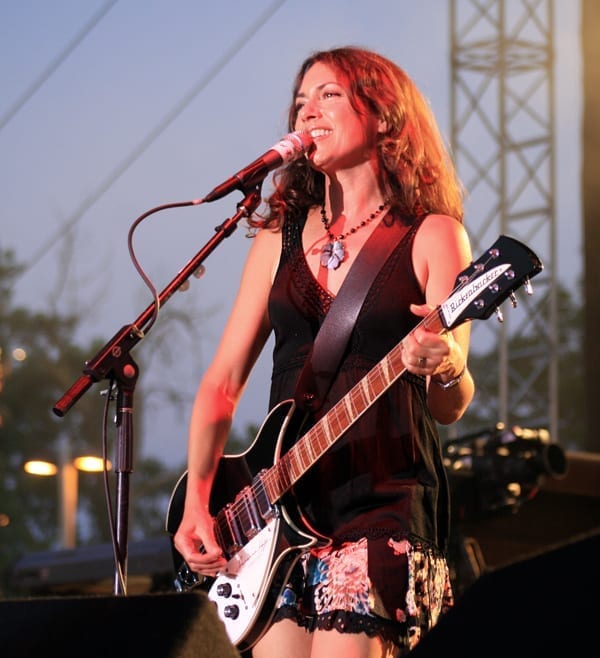 Susanna Hoffs, in a black dress, sings into a microphone while playing a black electric guitar at an outdoor evening show.