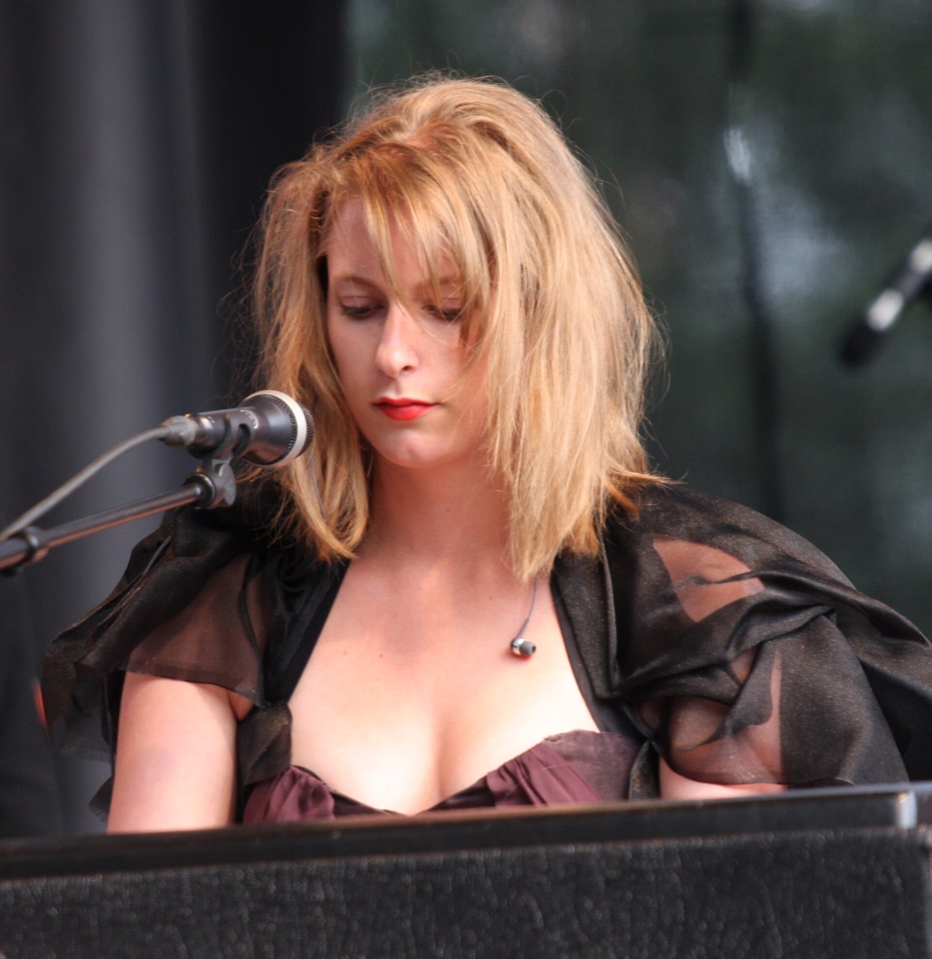 Susanne Sundfor, a young woman with tousled blonde hair and red lipstick, looks down while singing into a microphone on an outdoor stage, wearing a sheer black wrap over a purple dress.