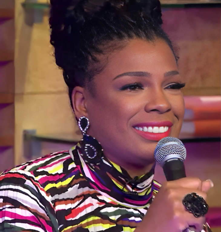 Syleena Johnson smiling and holding a microphone on a brightly lit television set, wearing large drop earrings and a multicoloured striped top