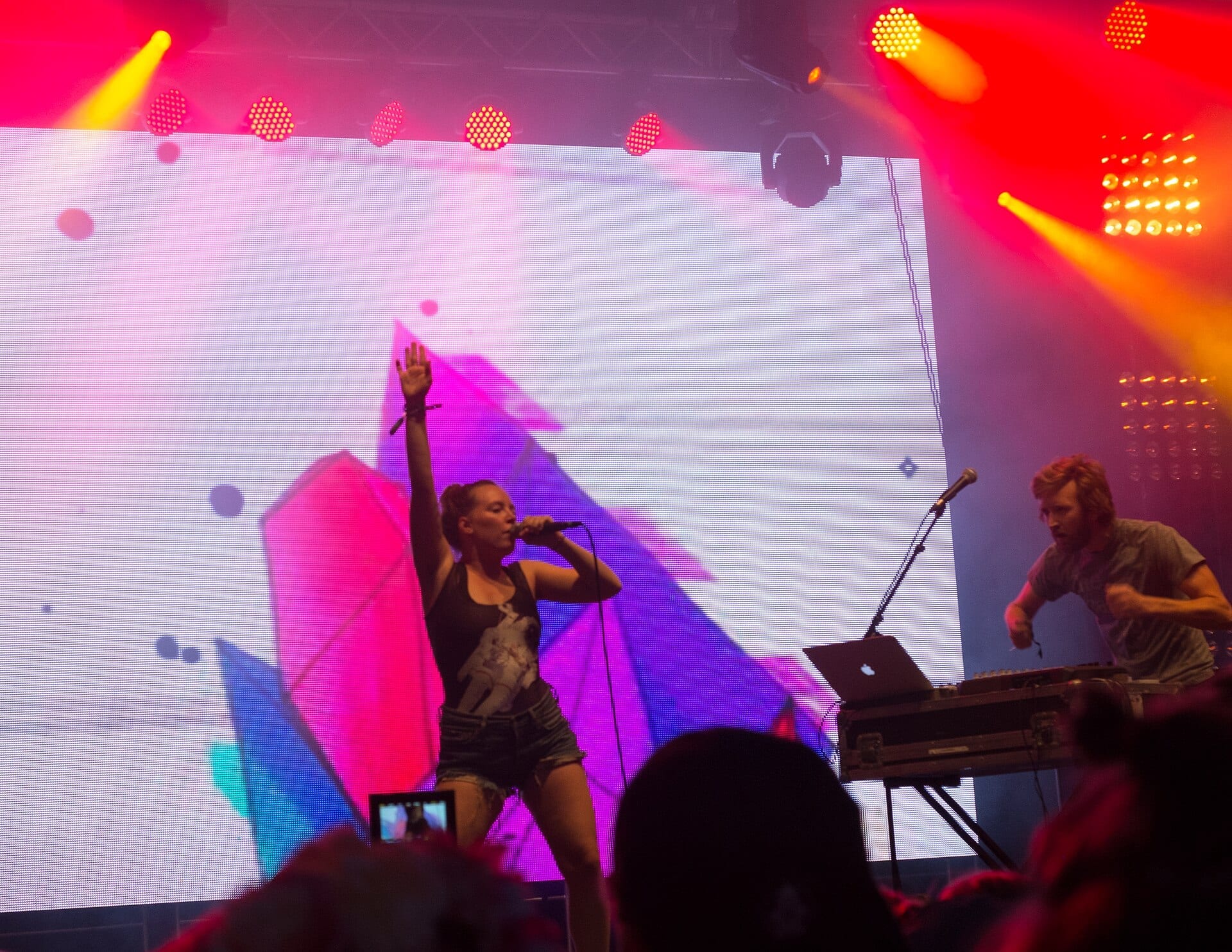 A singer raises one arm while singing into a microphone as her bandmate works over a laptop and controllers, both lit against a bright LED backdrop.