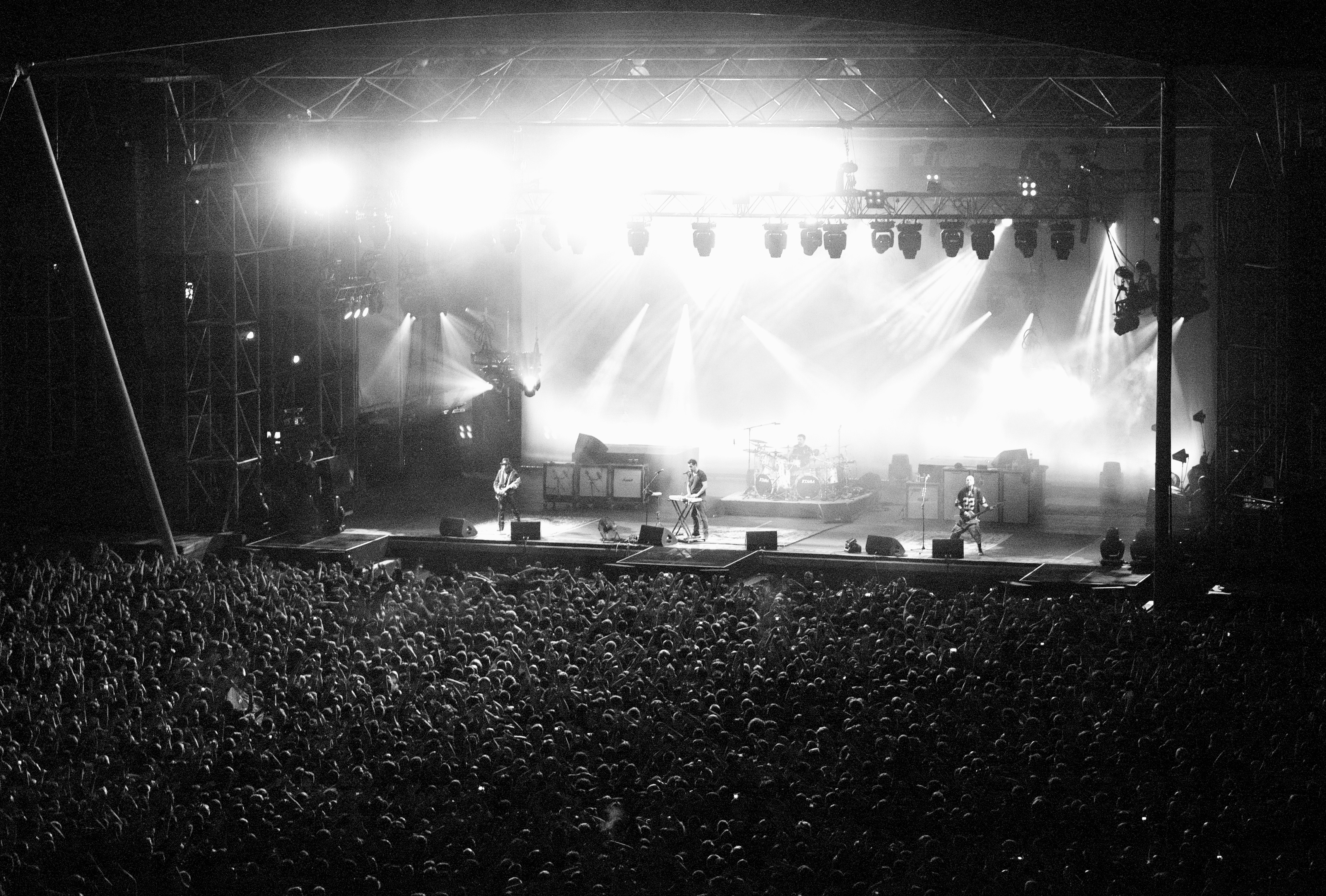 System of a Down play a large open-air stage in a black and white photograph, floodlights above them and a dense crowd filling the foreground.