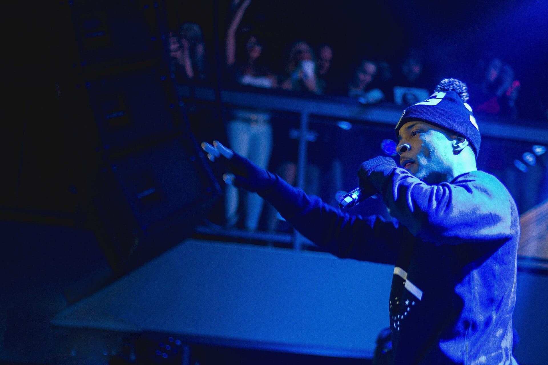T.I. performs on stage at night, pointing toward the crowd while holding a microphone, under blue stage lighting.