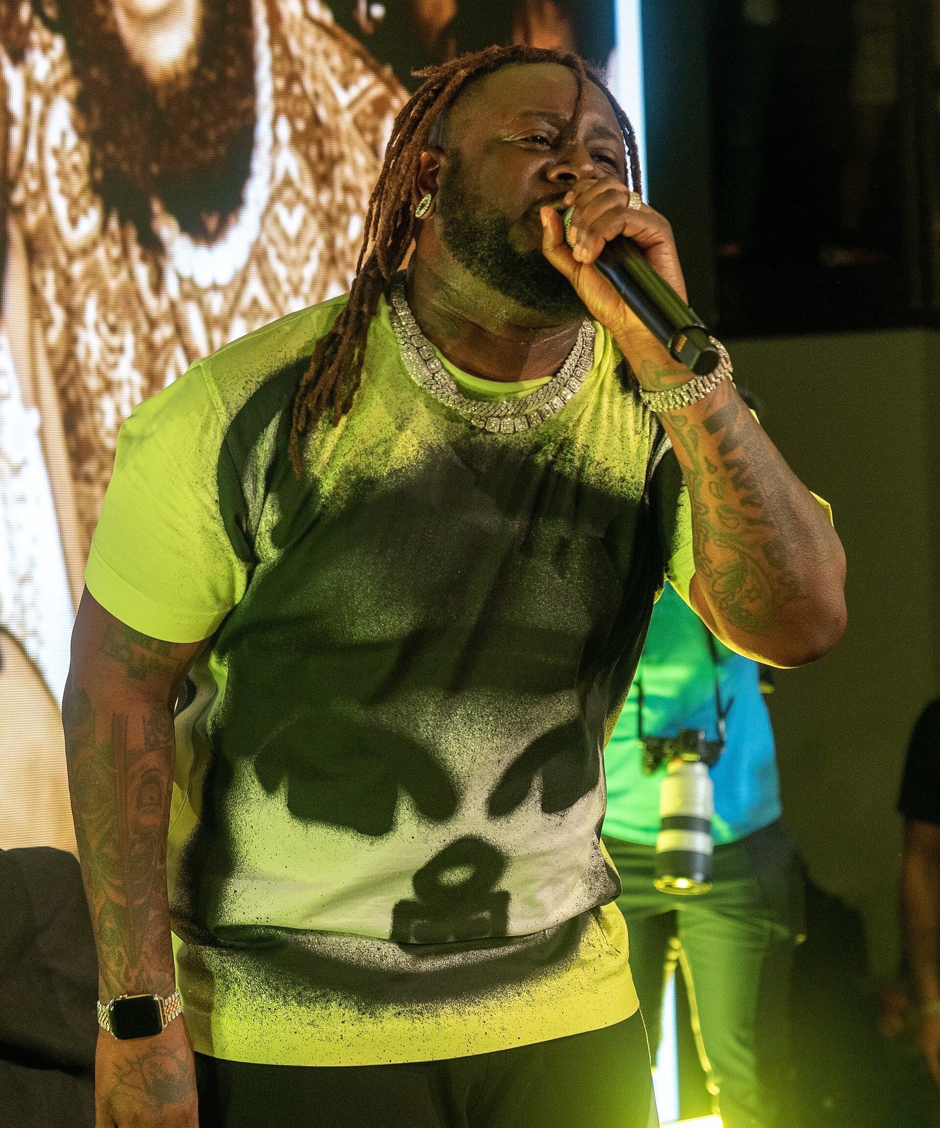 T-Pain sings into a microphone on stage, wearing a neon green tie-dye shirt and layered diamond chains.
