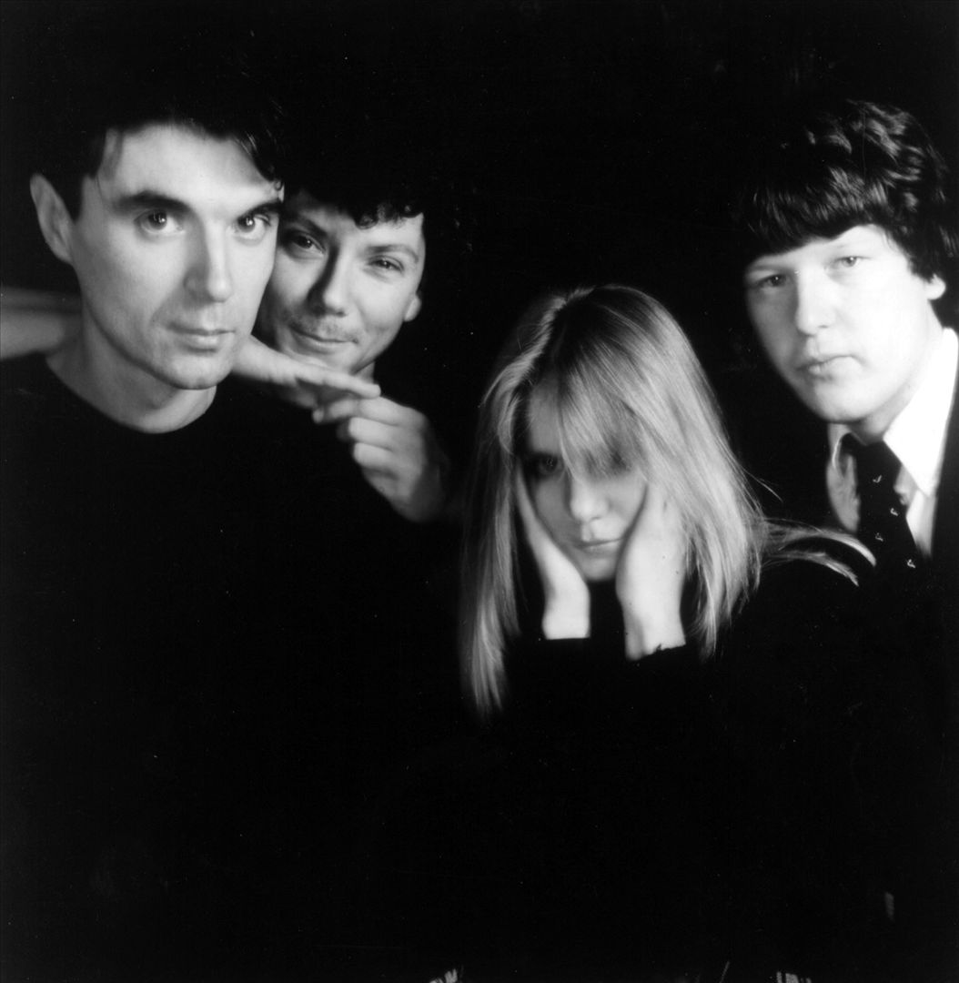 A black and white group portrait of the four members of Talking Heads against a dark background, David Byrne at left, Tina Weymouth holding her hands to her ears in the centre.