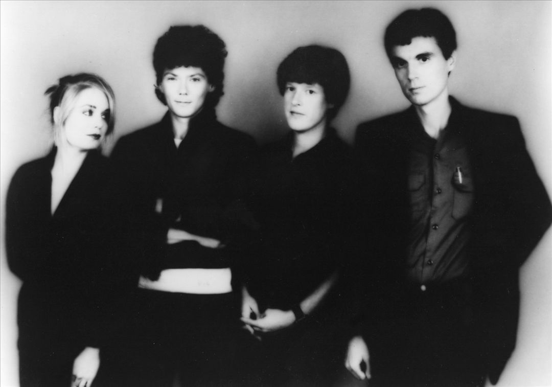 The four members of Talking Heads stand shoulder to shoulder in a black and white studio portrait, all in dark clothes against a plain pale wall.