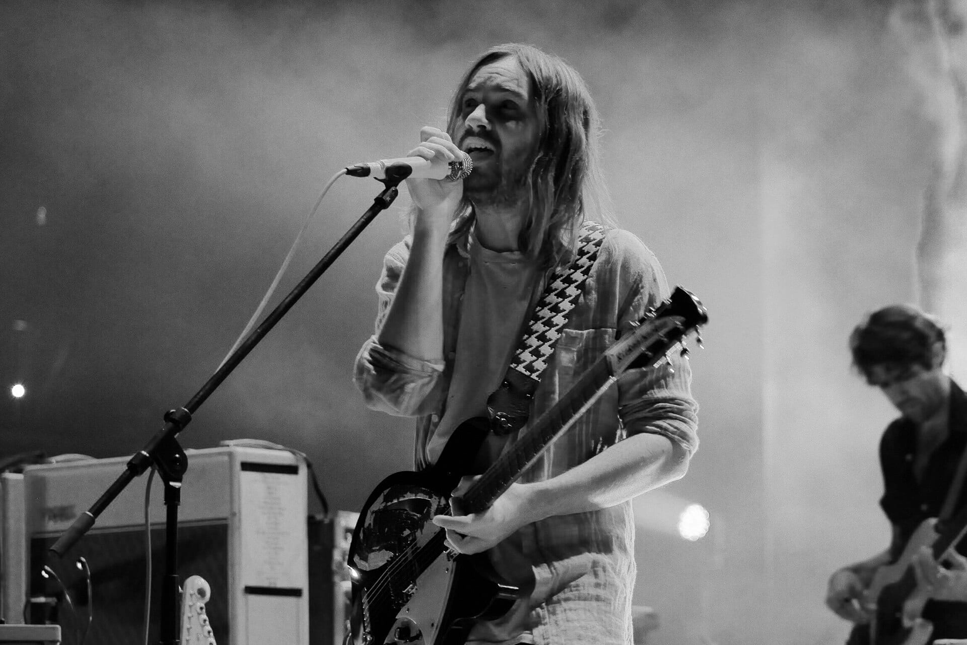 A long-haired singer plays an electric guitar and sings into a microphone in a black and white photo, a bandmate playing behind him.