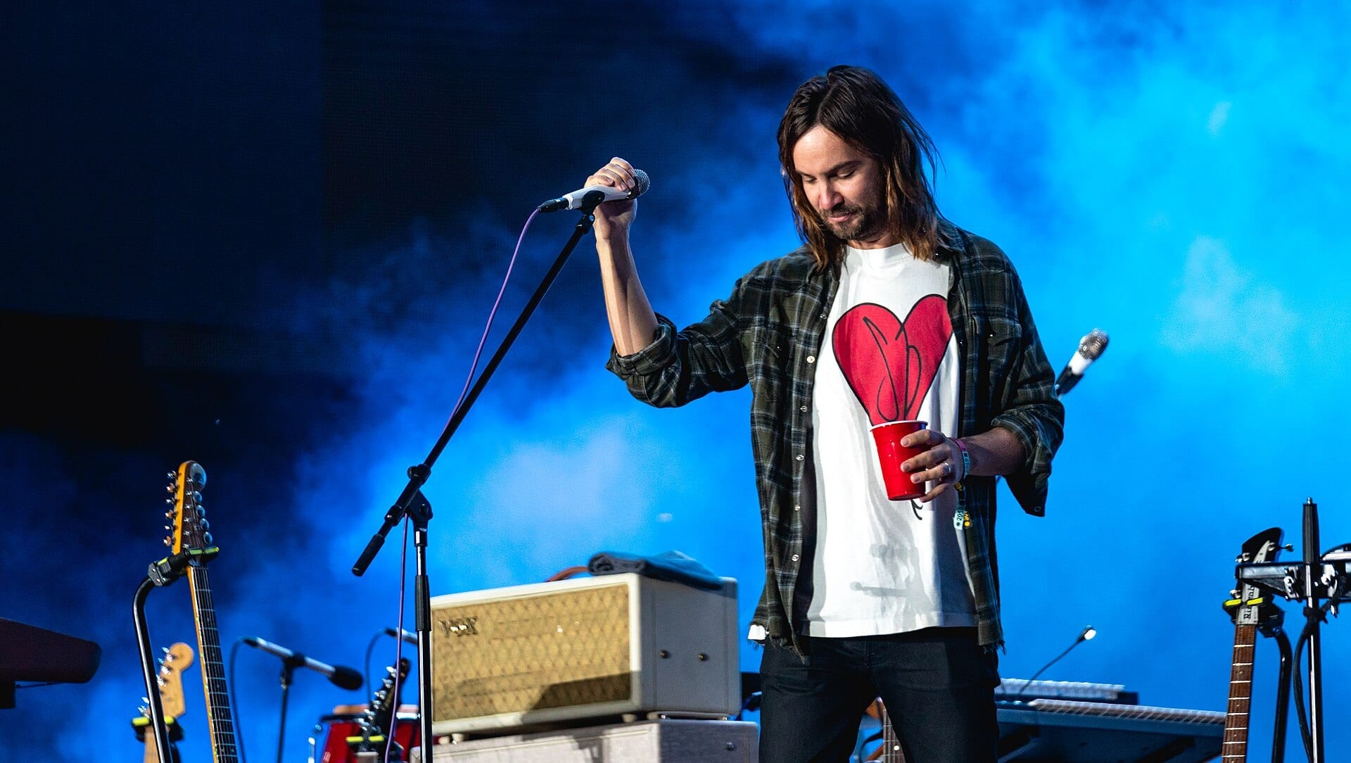A long-haired singer lifts a microphone off its stand and looks down, a red cup in his other hand, against blue stage haze.