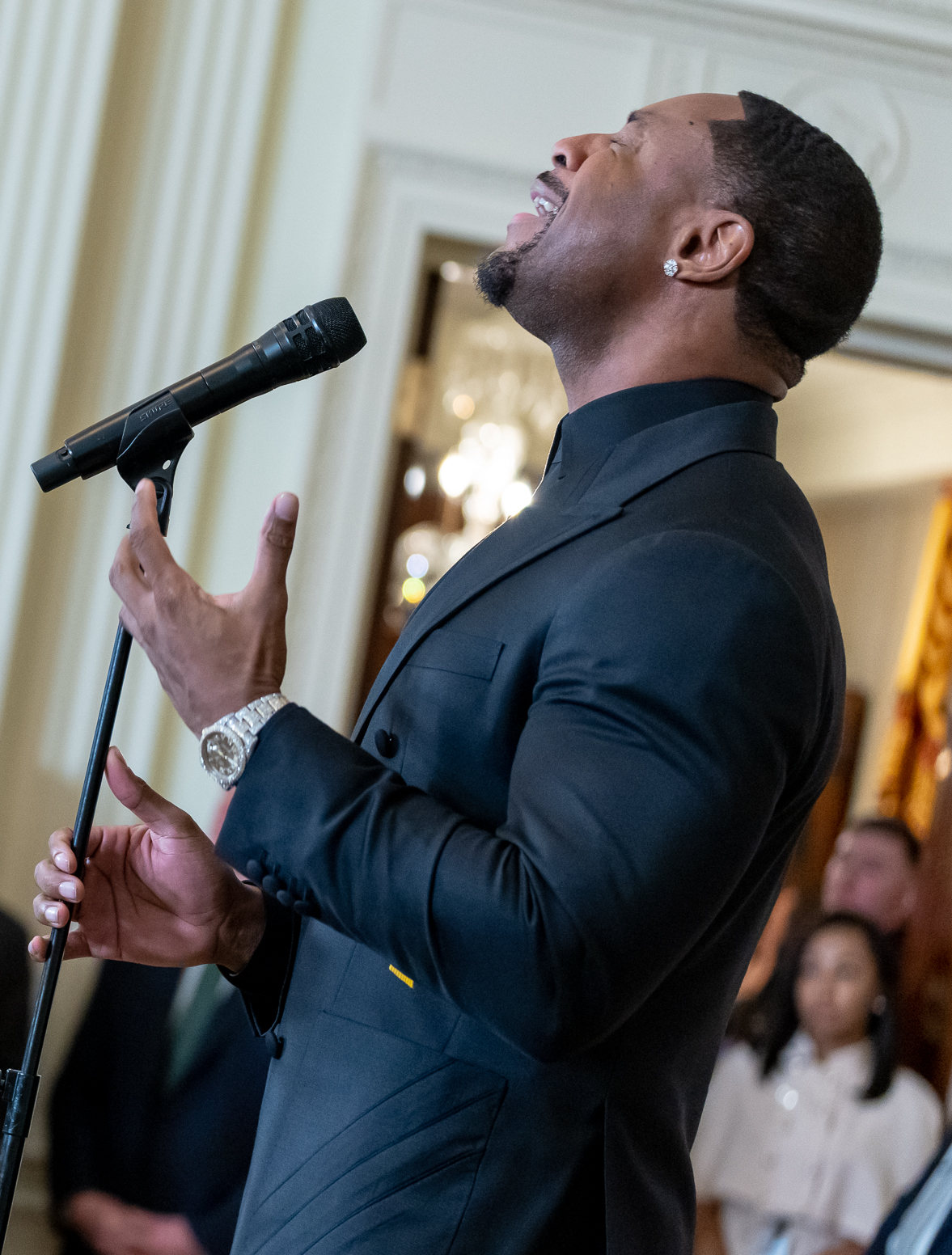Tank sings with his head tipped back and one hand raised beside a microphone stand, in a black suit and a heavy watch, in a pale panelled room.