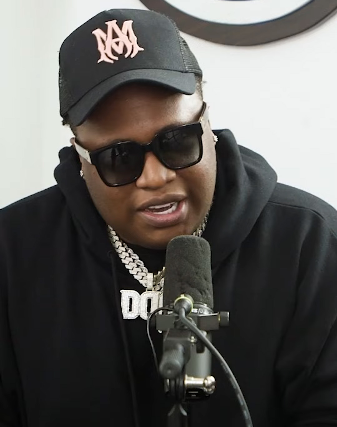 Tay Keith in a black cap and dark sunglasses, wearing a black hoodie and a diamond chain, talking into a studio microphone