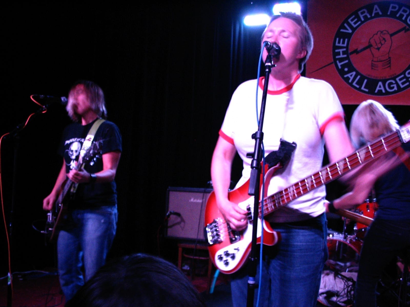 Two members of Team Dresch perform on a small club stage, playing guitars under stage lights in front of a red All Ages venue banner.