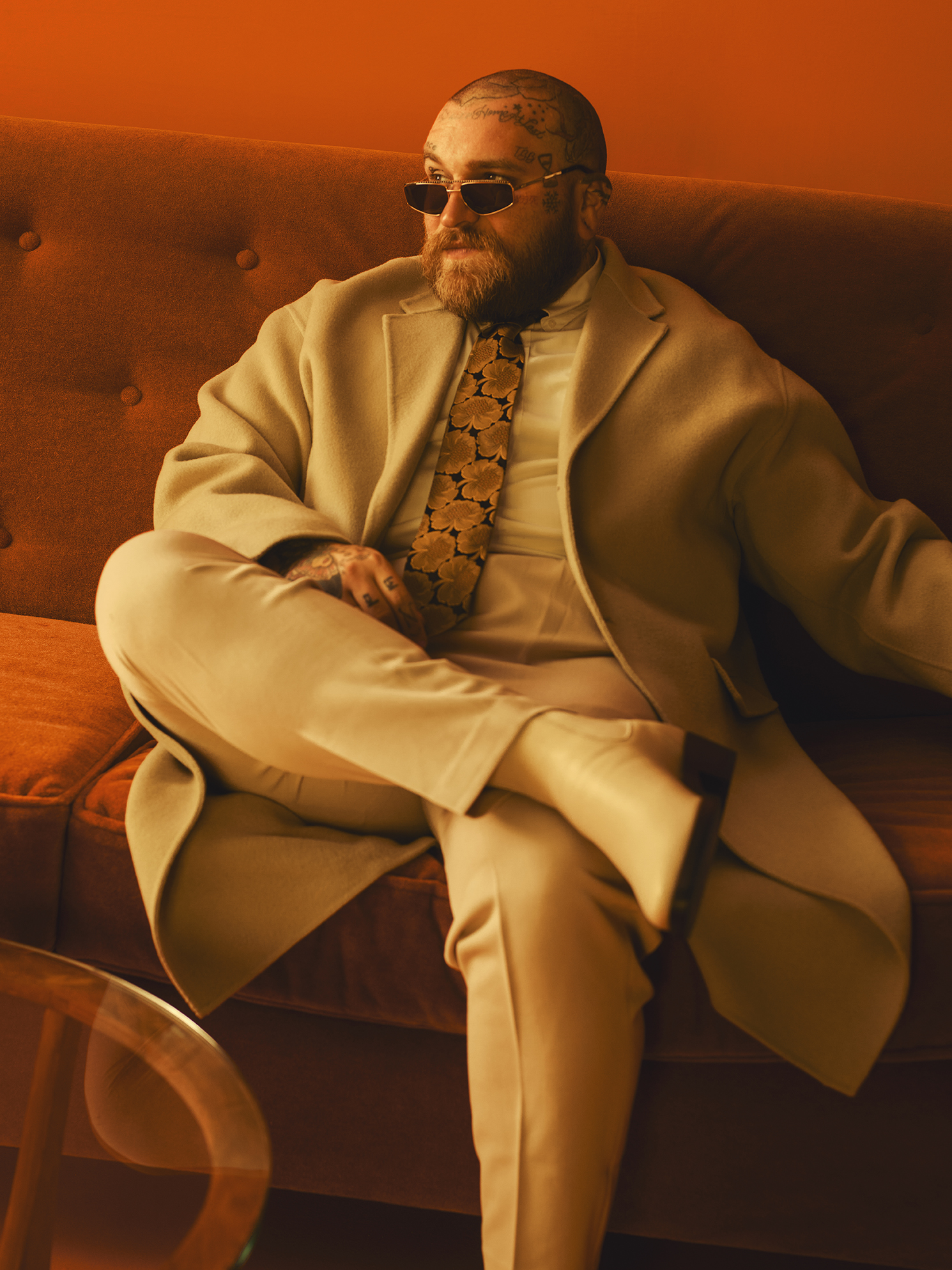 Teddy Swims sits back on a velvet sofa in a cream coat and suit with a floral tie, wearing narrow sunglasses, his head and face tattoos visible under warm orange light.