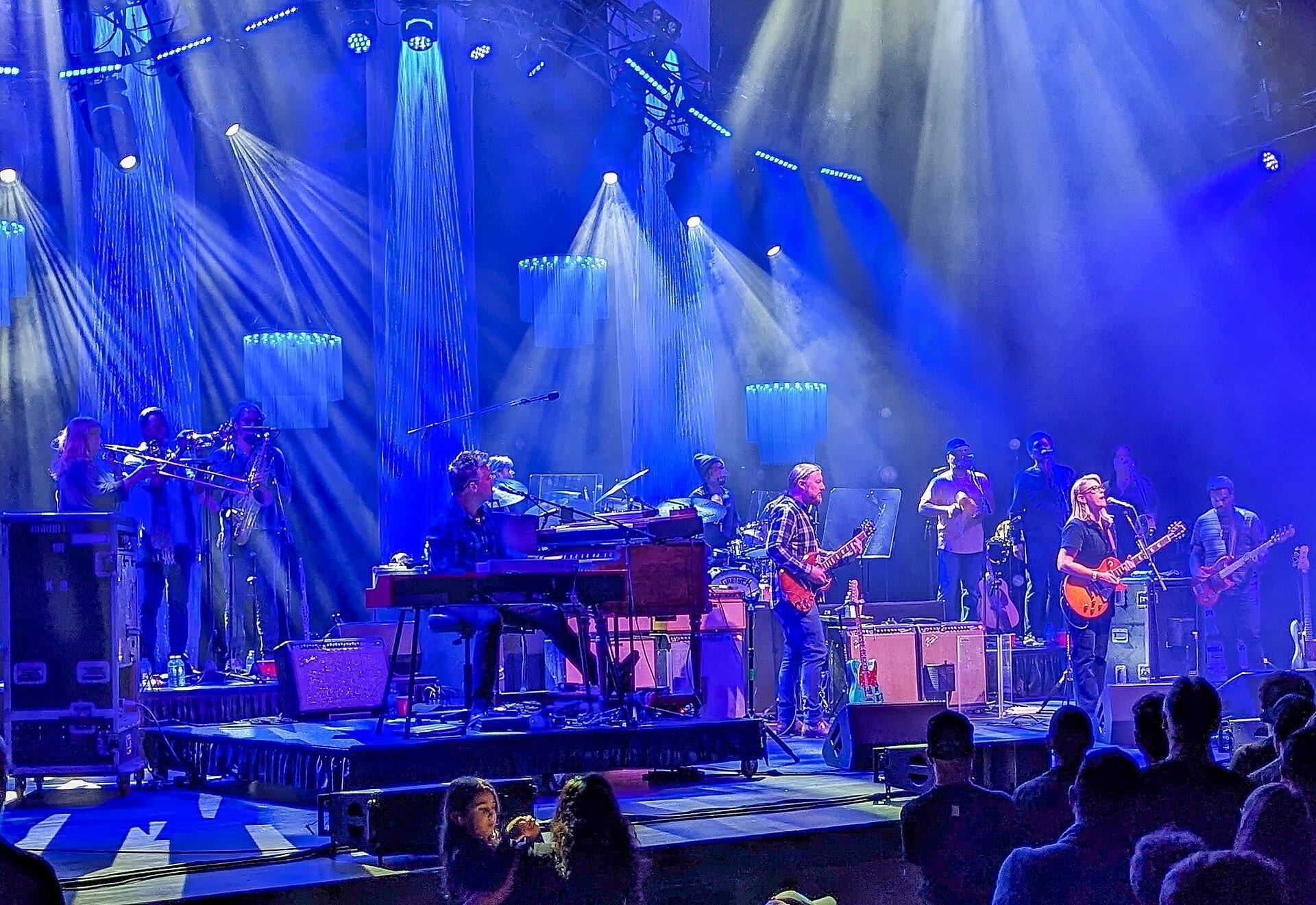 Tedeschi Trucks Band performs as a full ensemble on stage under blue lighting, with horn players, keyboards and multiple guitarists.