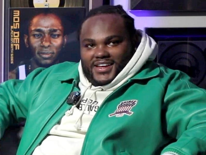 Tee Grizzley sits and speaks during an interview, wearing a green jacket over a cream hoodie, with a Mos Def album cover visible on the wall behind him.