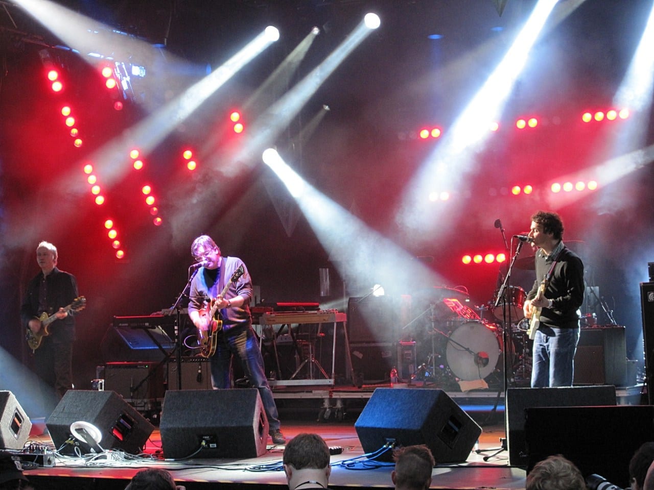 Three members of Teenage Fanclub play guitars on a stage crossed by white spotlight beams, with rows of red lamps behind them.