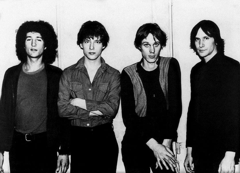 Four young men in dark clothes stand shoulder to shoulder against a plain wall in a black and white publicity photo.