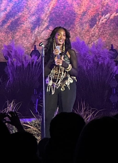 Tems, wearing layered gold necklaces over a black outfit, sings into a microphone on a stage backed by purple-lit foliage.