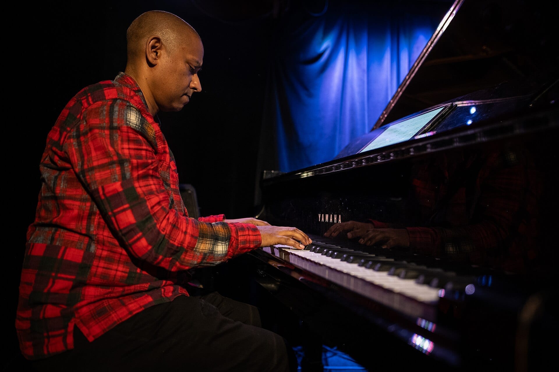 Terence Collie plays a grand piano in profile under blue stage light, wearing a red plaid flannel shirt, with sheet music displayed on a tablet propped on the piano.