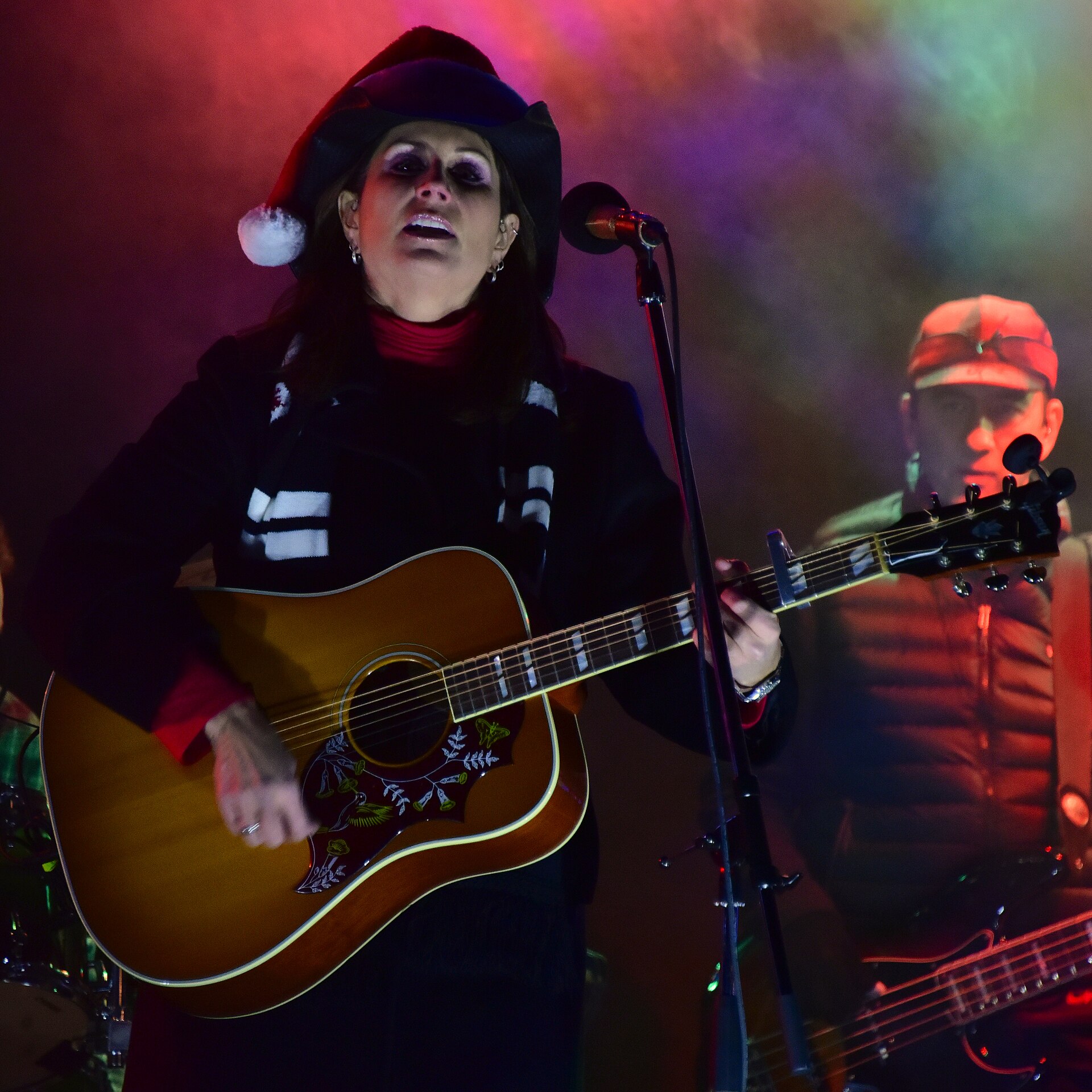Terri Clark sings at a microphone playing an acoustic guitar, wearing a black cowboy hat trimmed like a Santa hat and a heavy dark coat, a bass player behind her.