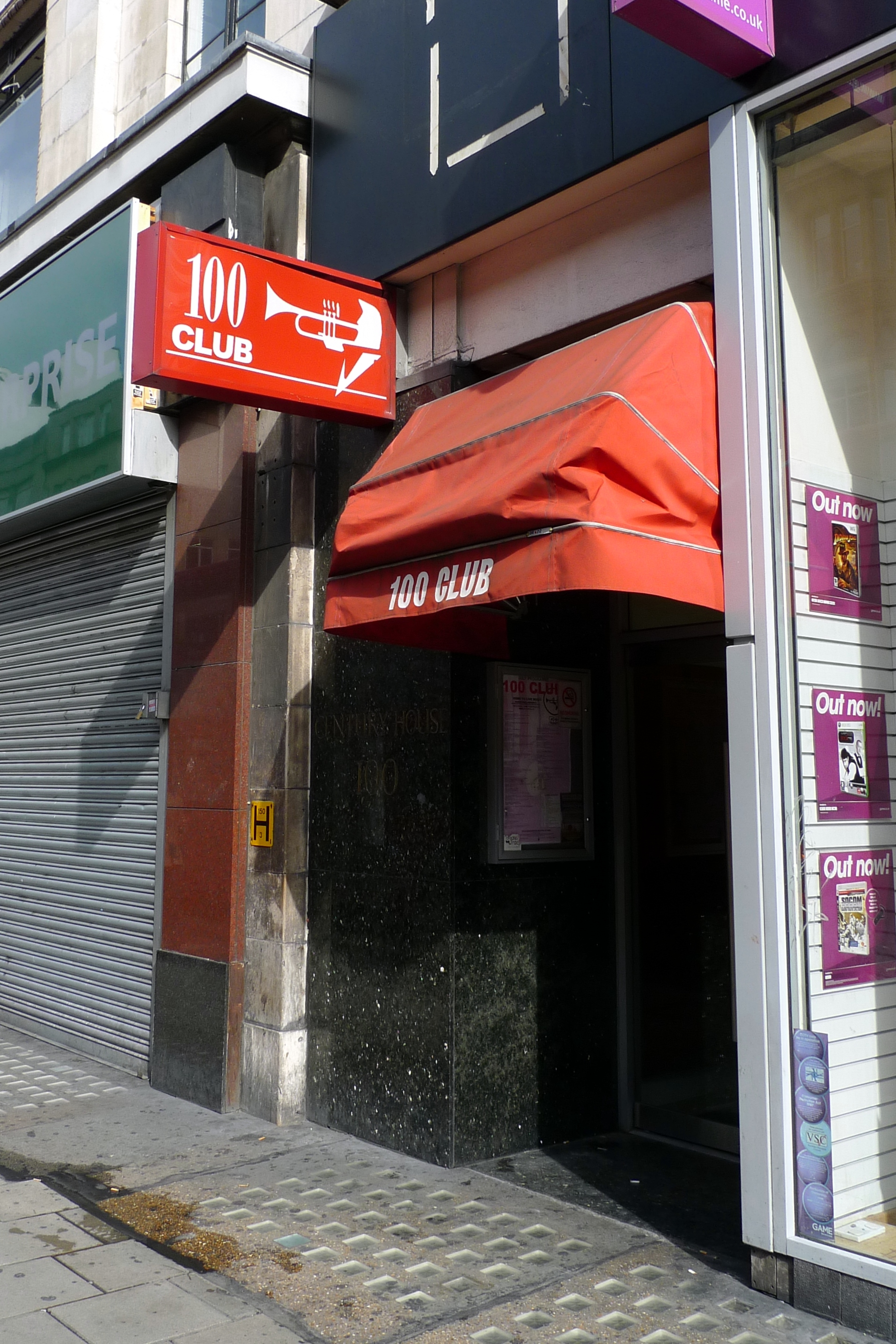 The 100 Club shopfront in daylight, red trumpet sign and orange awning wedged between a shuttered unit and a games shop window.