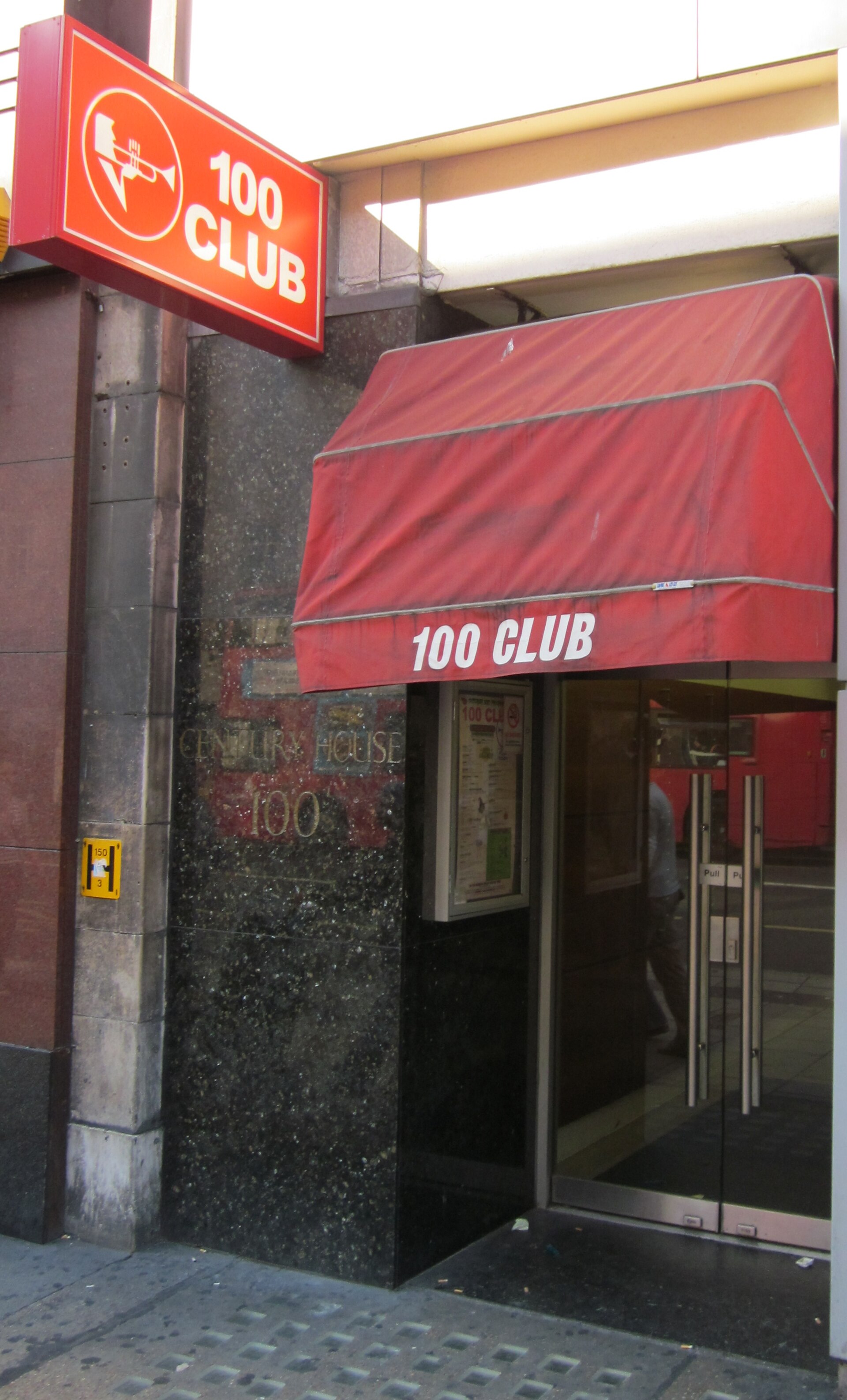 The 100 Club entrance on Oxford Street, a red hanging sign with a trumpet logo above a red awning and a basement stairwell.