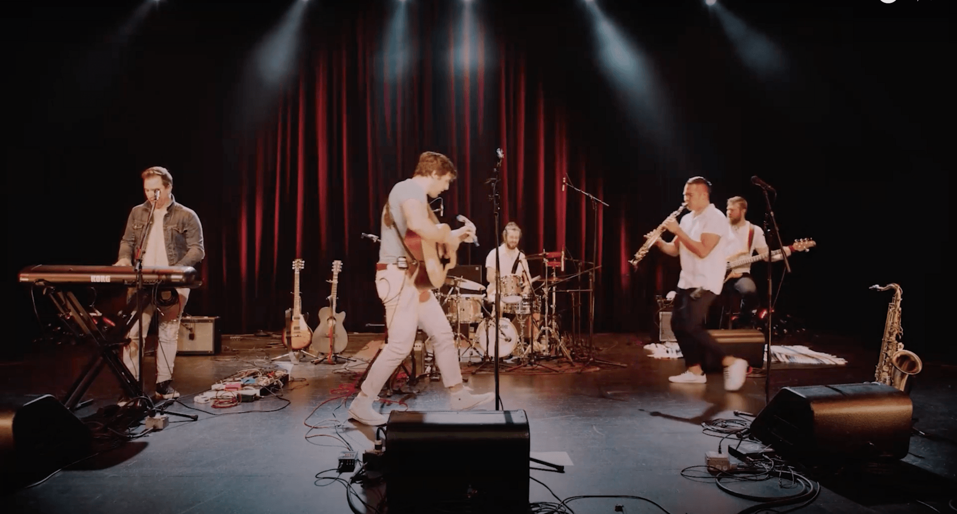 The 502s perform on a stage with a red curtain backdrop, band members playing keyboard, acoustic guitar, drums, saxophone, and electric guitar.