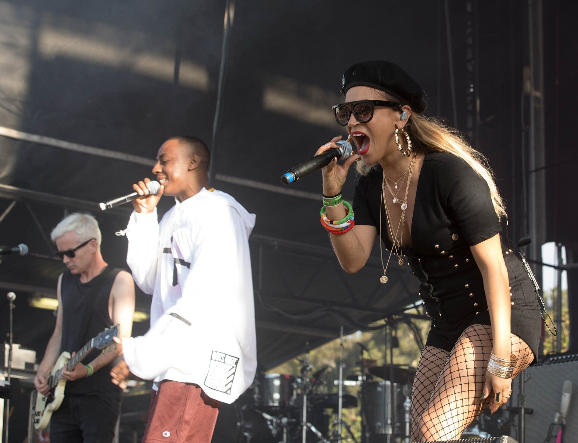 Three members of The Avalanches perform on an outdoor festival stage in daylight: a guitarist and a vocalist in a white hoodie sing into microphones on the left, while a vocalist in a black studded outfit and sunglasses sings into a microphone in the foreground.