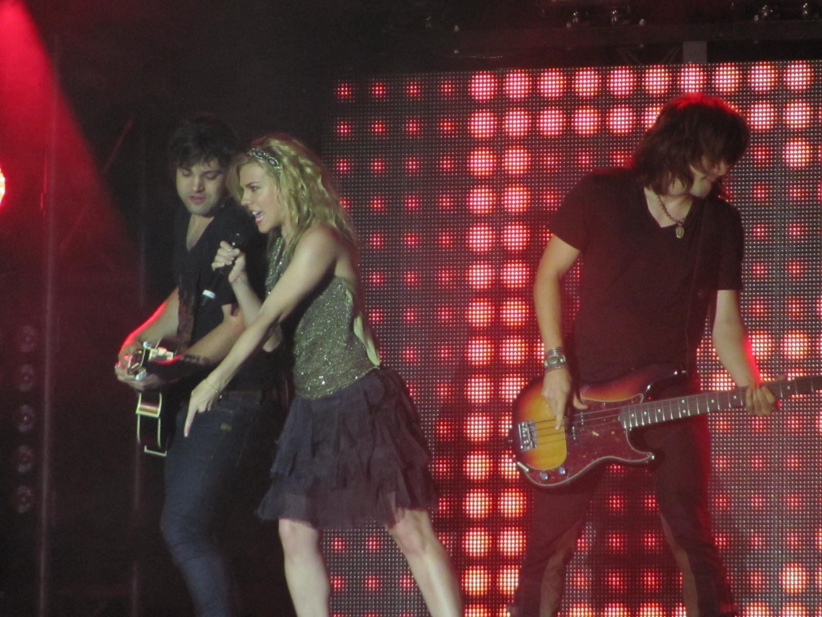 The Band Perry's three original members perform on a red-lit stage: a singer at center with a handheld microphone, a guitarist at left, and a bassist at right.