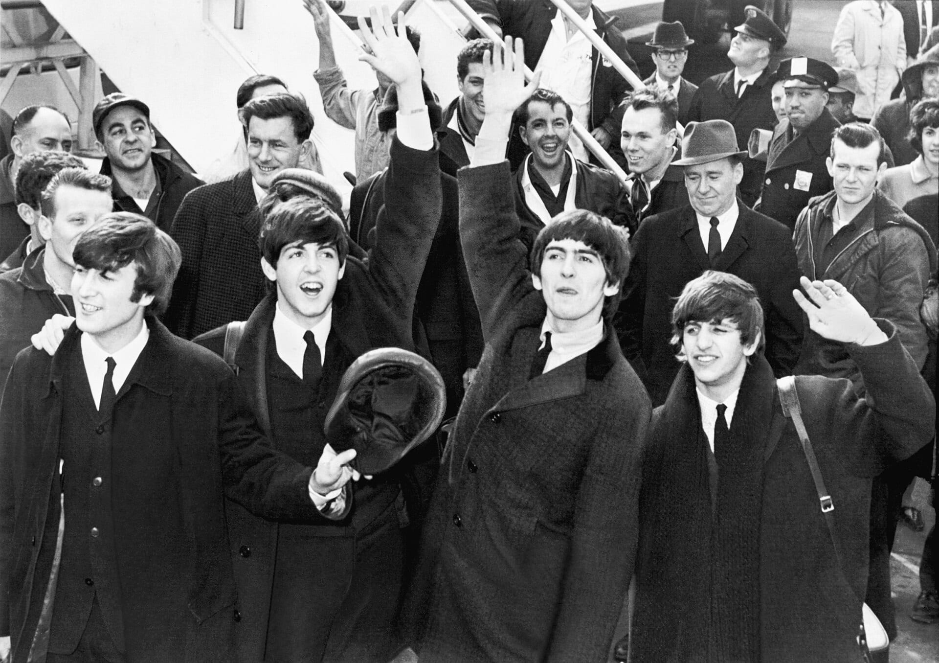 The four members of The Beatles smile and wave while descending an airplane staircase, surrounded by police officers and airport staff.