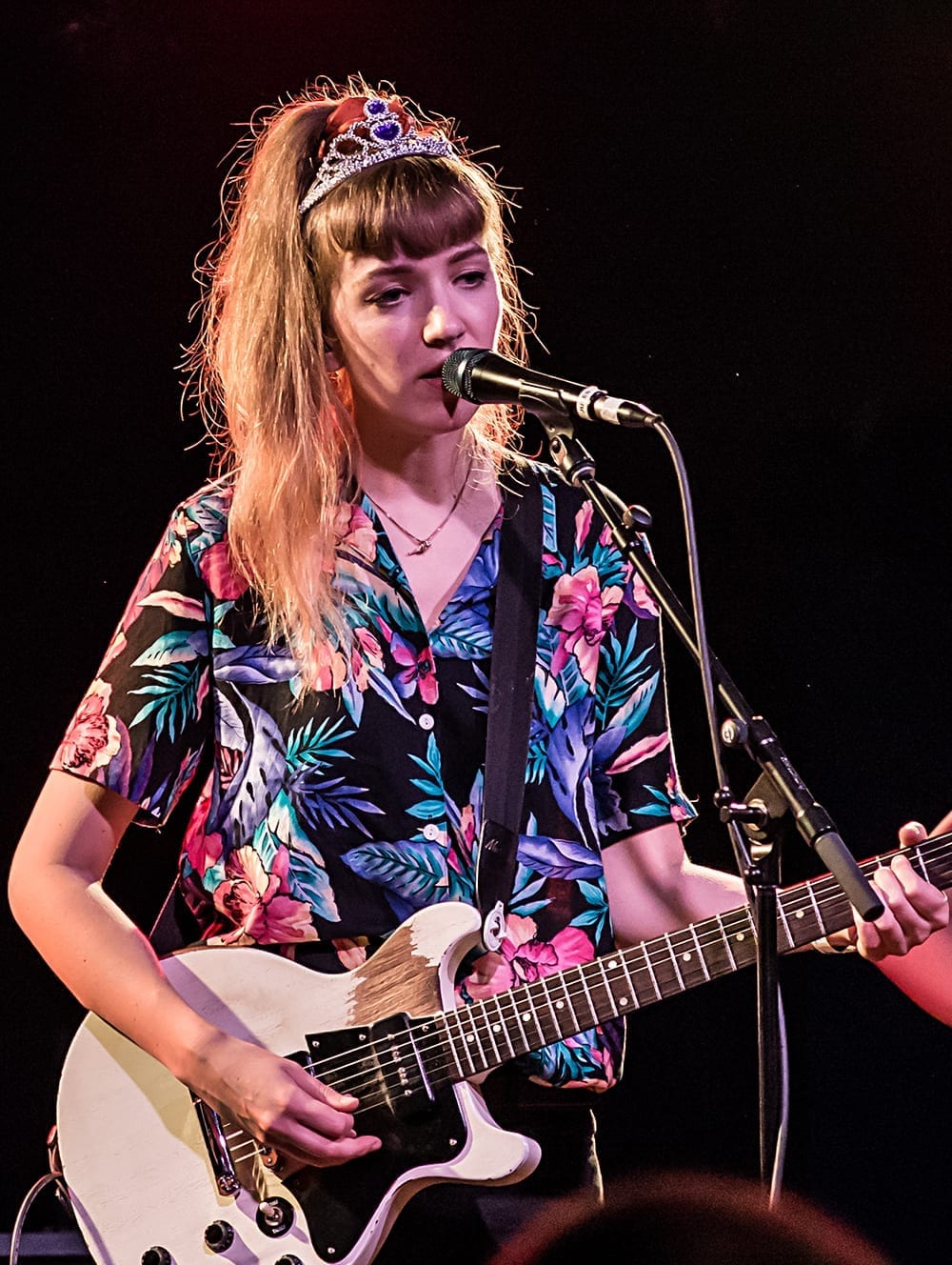 A singer from The Big Moon, wearing a tiara and a floral shirt, sings into a microphone while playing a white electric guitar.