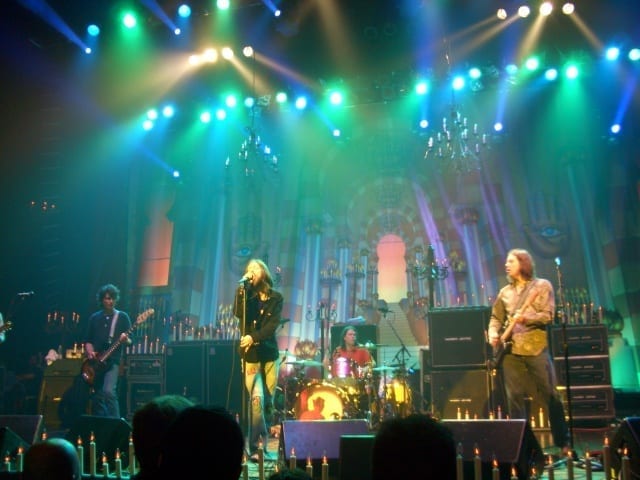The Black Crowes perform under green and blue stage lights on an ornate theater stage lined with candle-shaped lamps and chandeliers.
