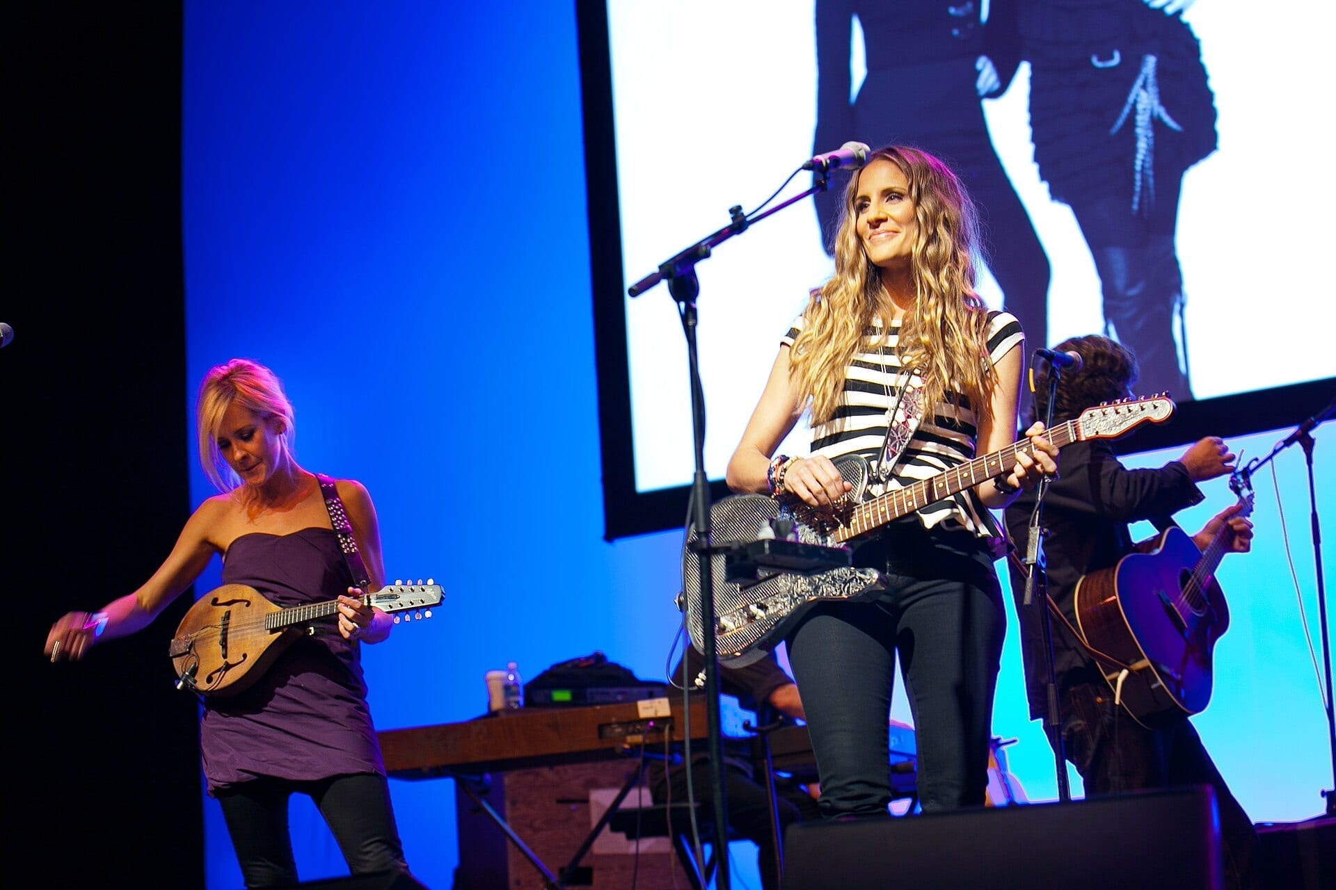 Two members of The Chicks perform on stage, the singer at center playing an electric guitar in a black and white striped top while a bandmate plays mandolin beside her.
