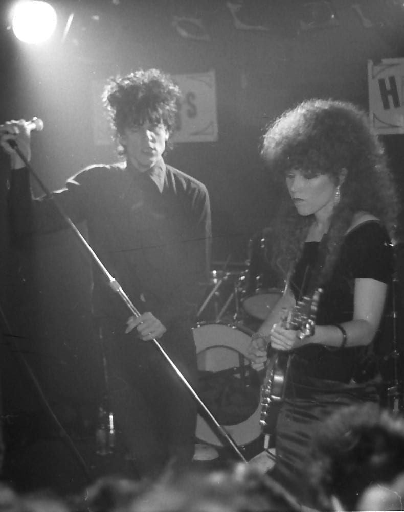 Black and white photo of Lux Interior gripping a microphone stand beside Poison Ivy, who plays guitar in a dark club.