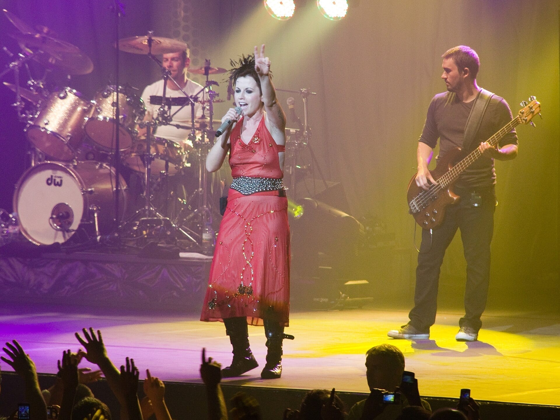 Dolores O'Riordan sings into a microphone with a raised hand in a red dress and black boots, flanked by a drummer and a bassist on a purple- and yellow-lit stage.