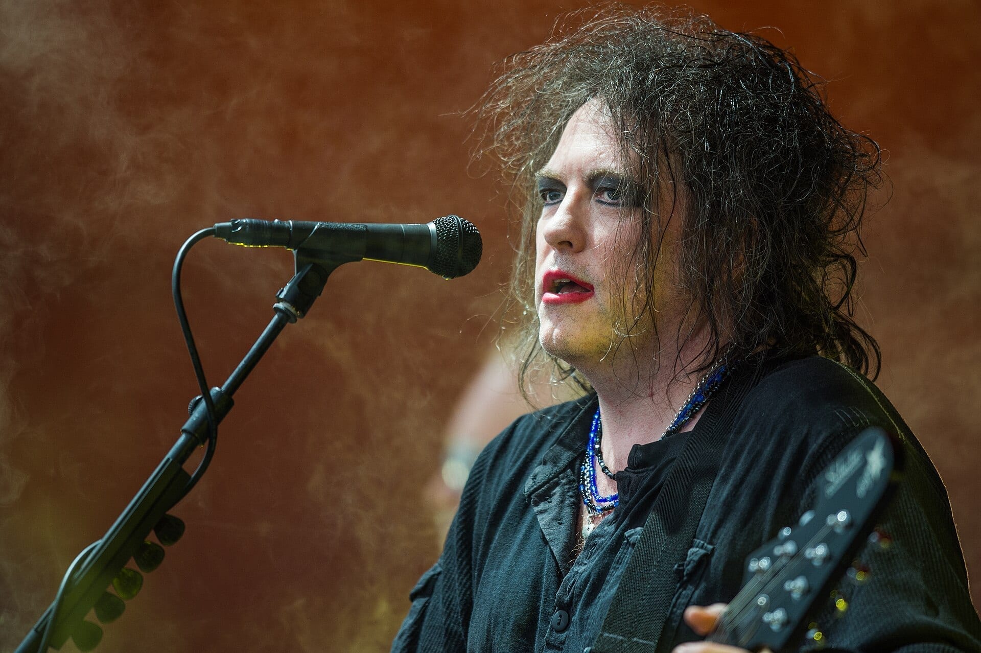 Robert Smith of The Cure sings into a microphone in close-up, his dark hair teased out, wearing smudged red lipstick and dark eye makeup with a black jacket and layered necklaces.