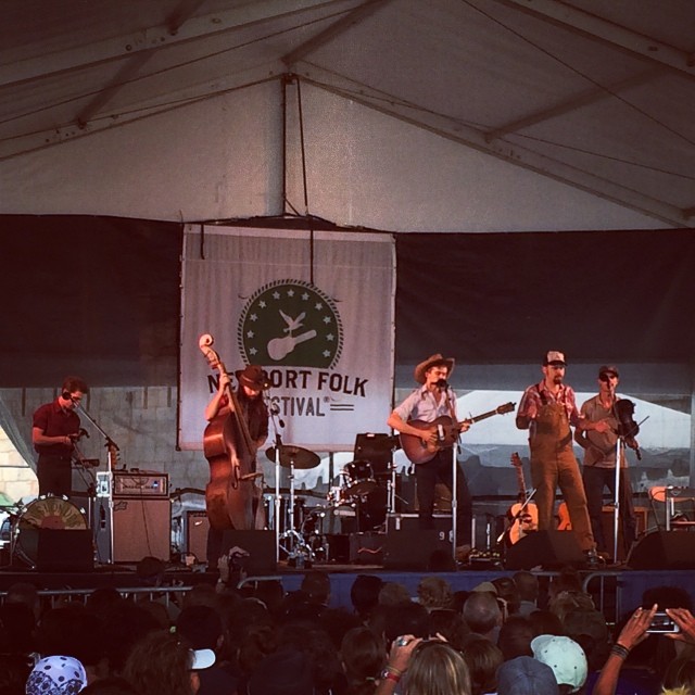 The Deslondes play an outdoor festival stage as a five-piece, upright bass on the left, two guitarists at the microphones and a fiddle player to the right.