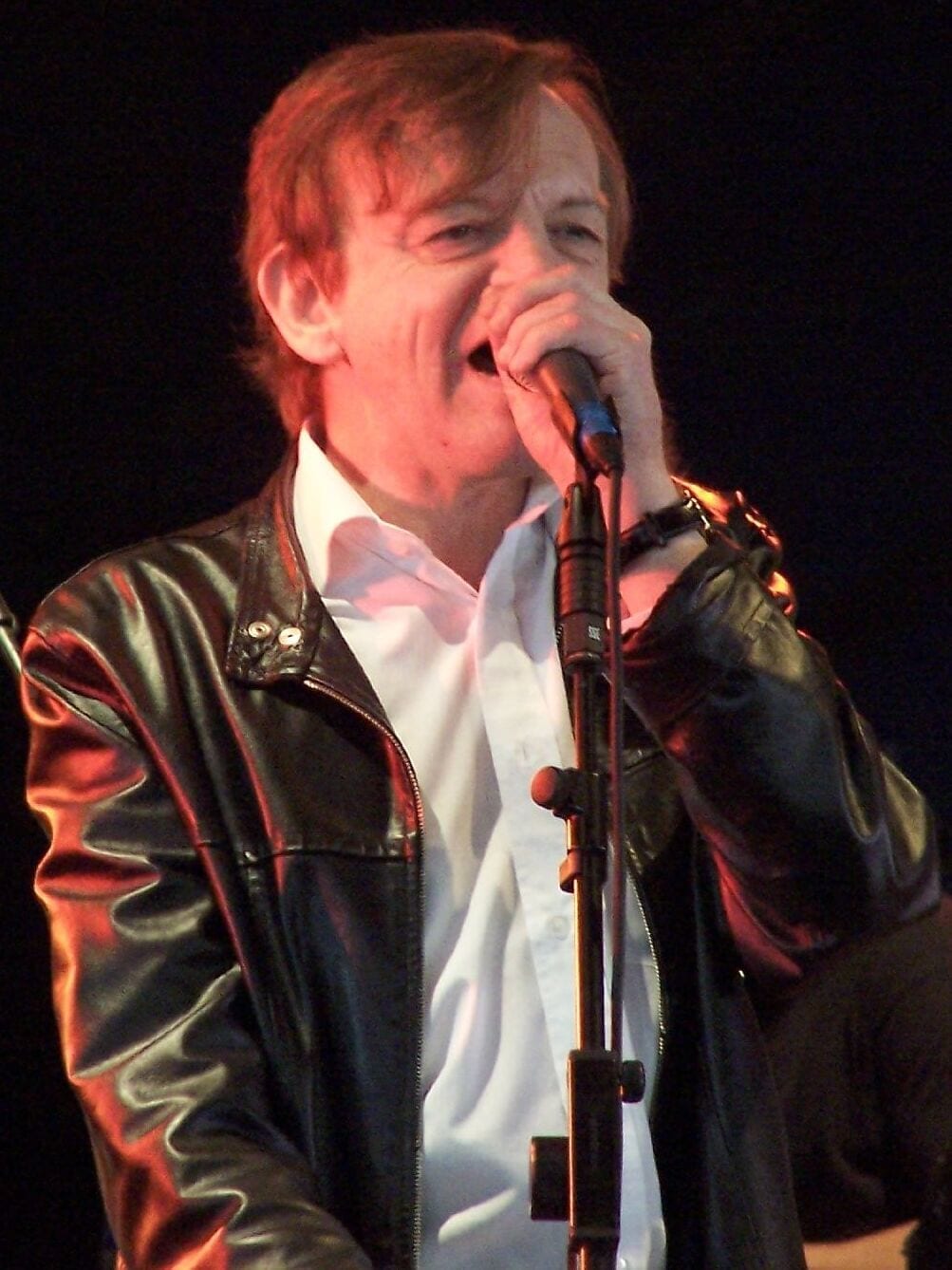 Mark E. Smith of The Fall sings into a handheld microphone on stage, wearing a black leather jacket over a white shirt.