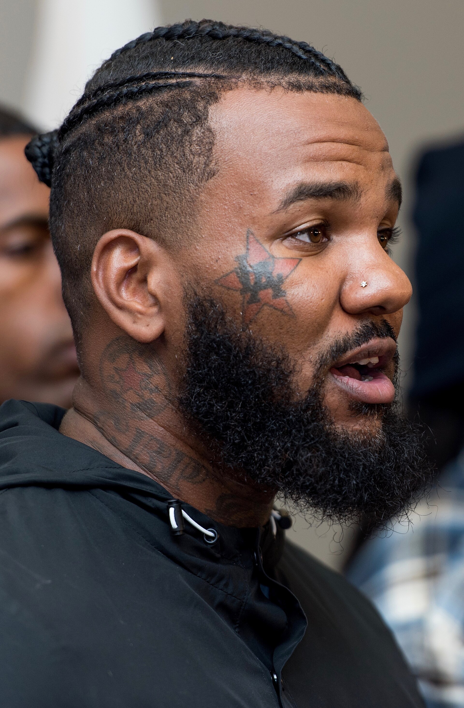 The Game speaks at a lectern in a black jacket and cap, the butterfly tattoo across his face visible, other speakers behind him.