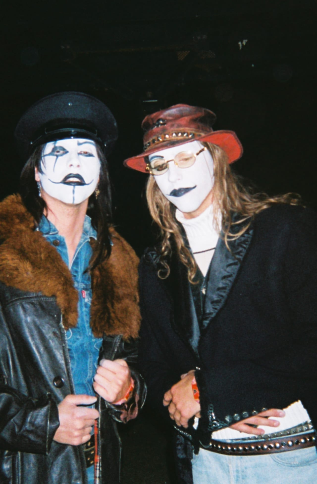 The two members of The Garden pose in white kabuki-style face paint, one in a fur-collared leather coat and cap, the other in a red hat and round glasses.