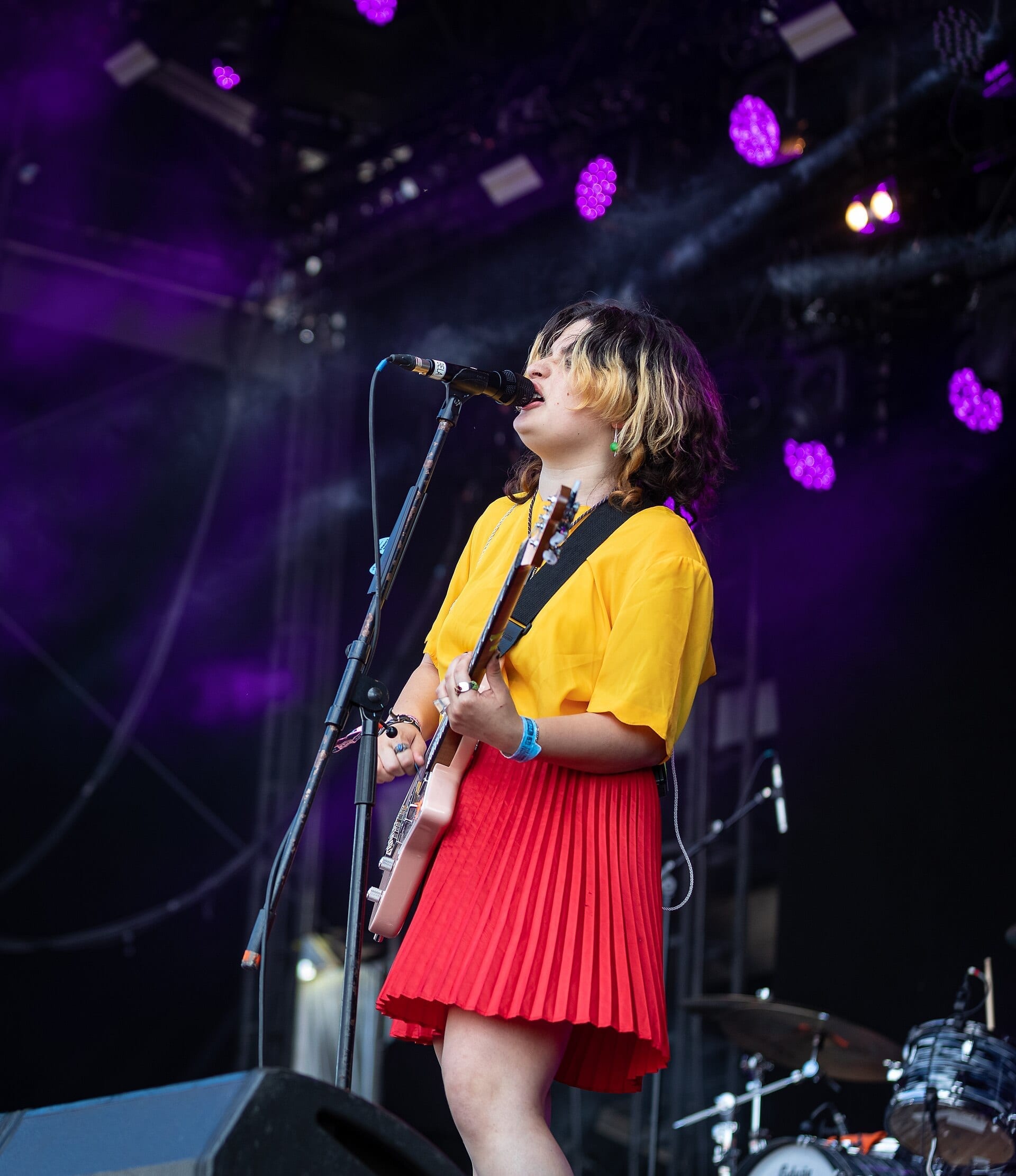 A member of The Linda Lindas sings into a microphone while playing guitar on an outdoor festival stage, wearing a yellow top and red pleated skirt.