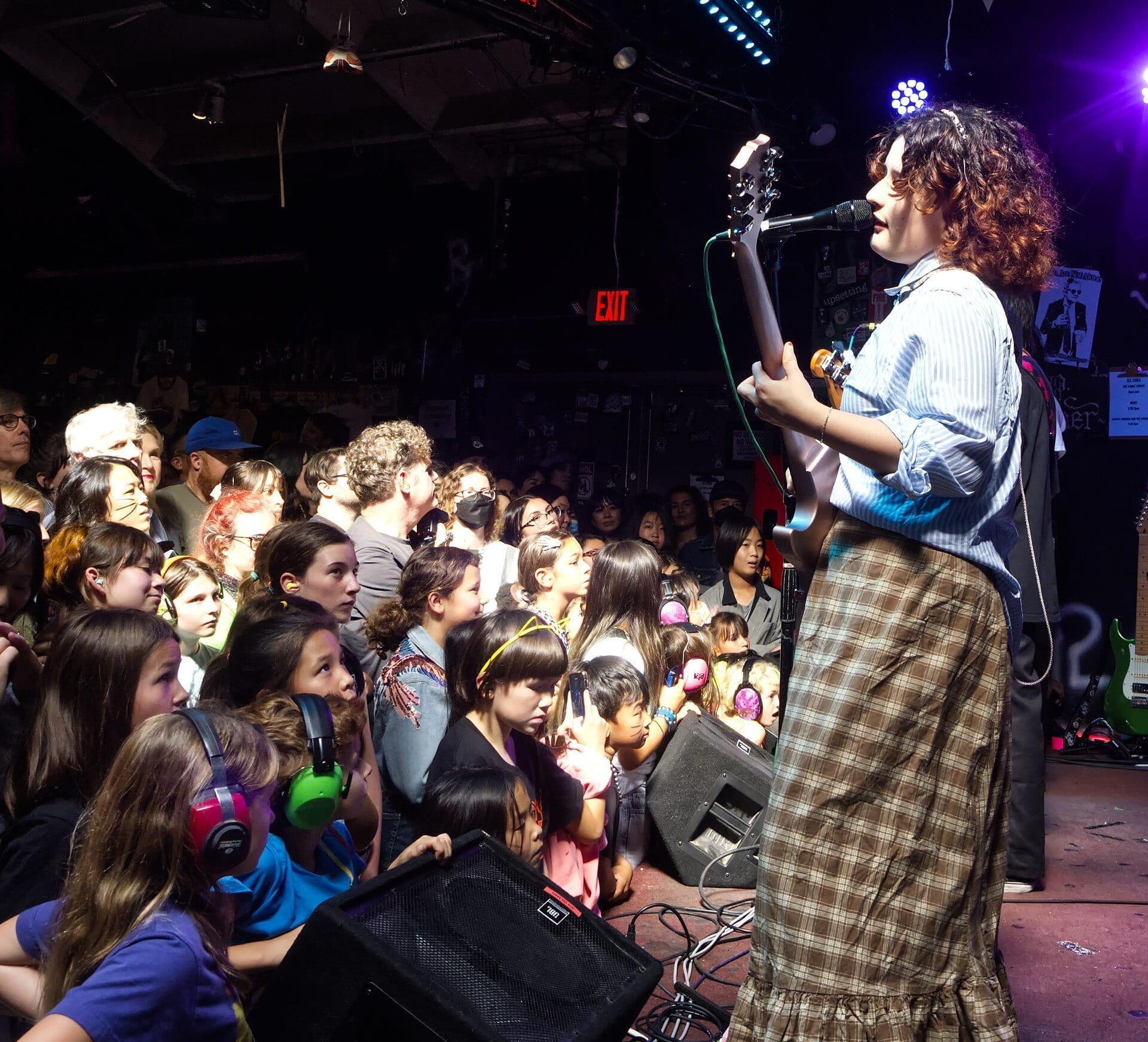 A member of The Linda Lindas sings and plays guitar at the edge of a low stage, facing a packed crowd of young fans, several of them in ear defenders.