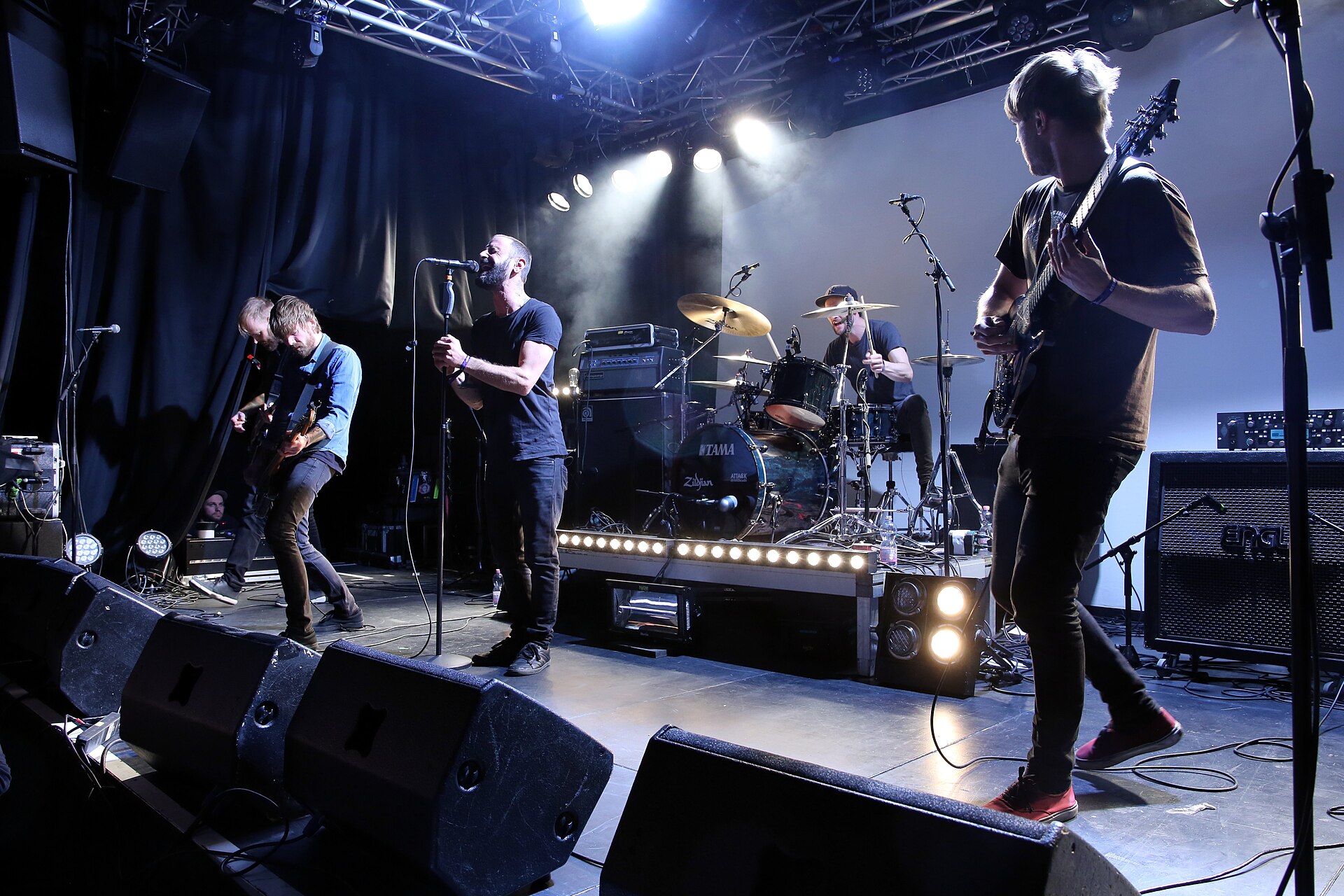 The Ocean play a small club stage, the singer centre at a microphone stand in a black t-shirt, guitarists either side and a drummer on a riser behind under white spotlights.