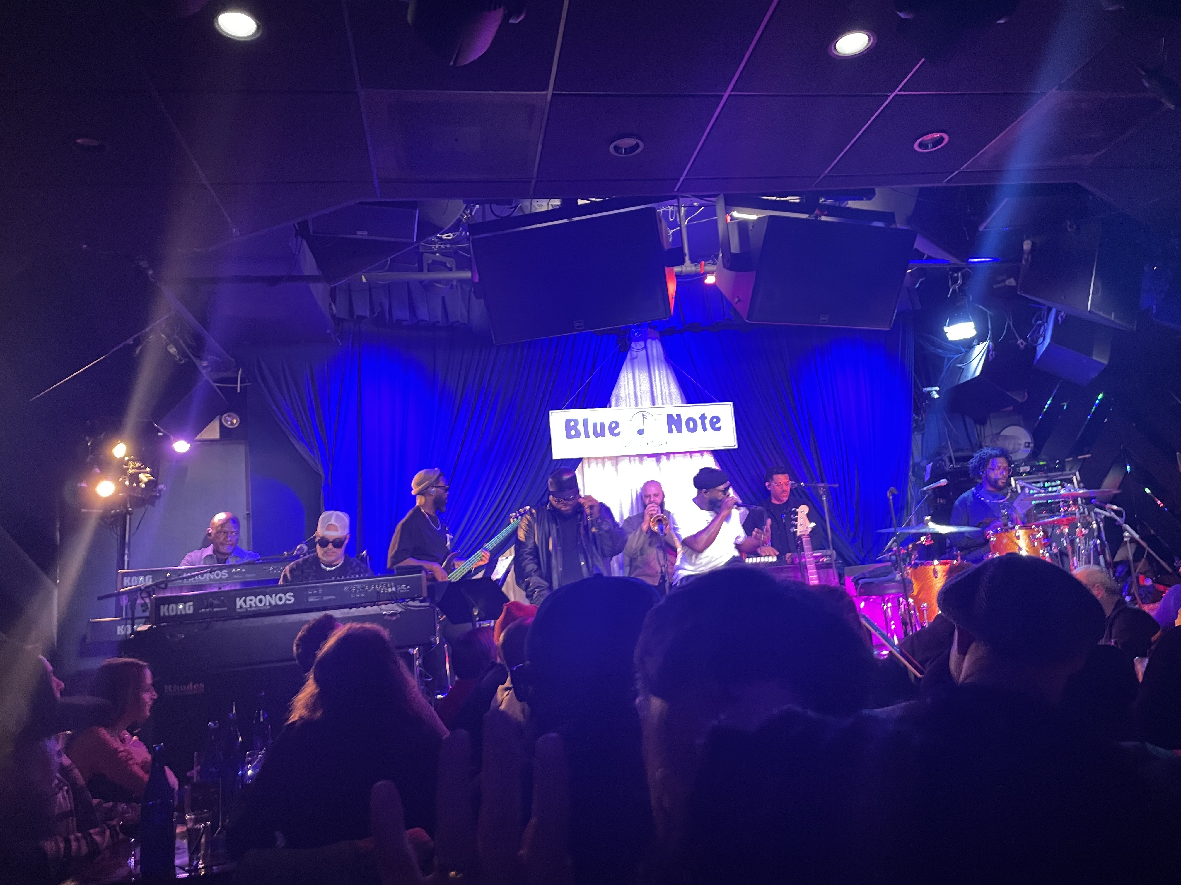 The Roots play a small club stage lit blue beneath a Blue Note sign, keyboards at the left and drums at the right, seen over the heads of the audience.